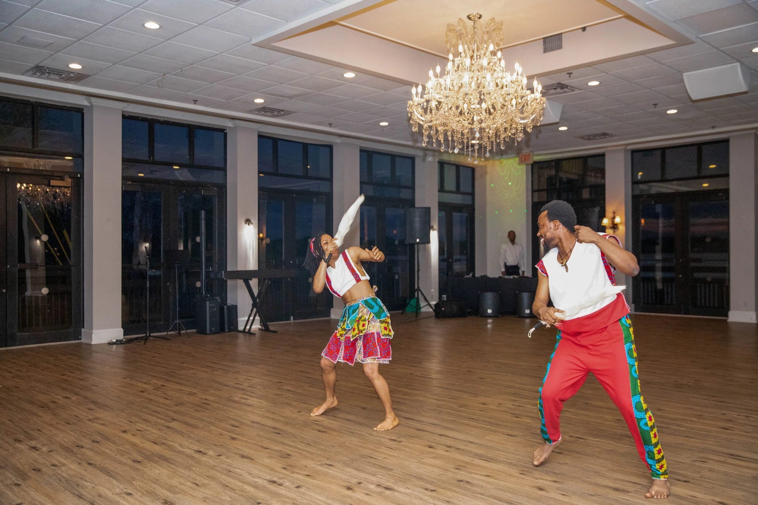 Two people dancing indoors at a celebration, dressed in colorful African attire, with a chandelier hanging from the ceiling.