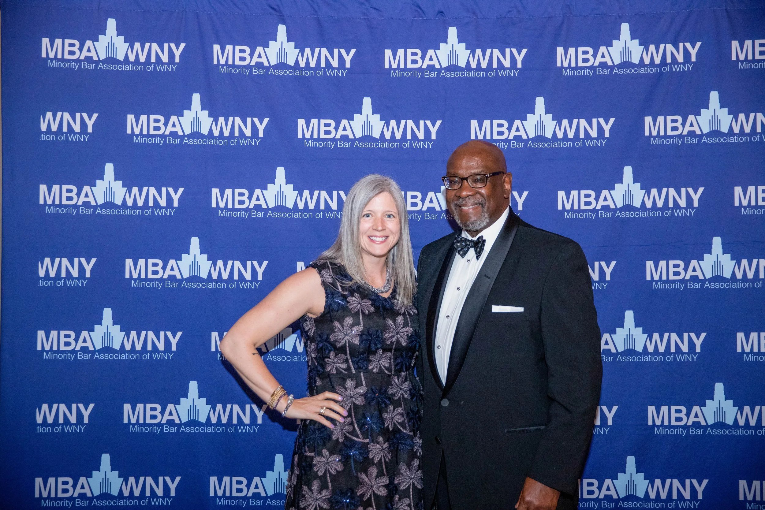 Two people, a woman and a man, dressed in formal attire, standing in front of a blue backdrop with the logo and text of the Minority Bar Association of WNY.