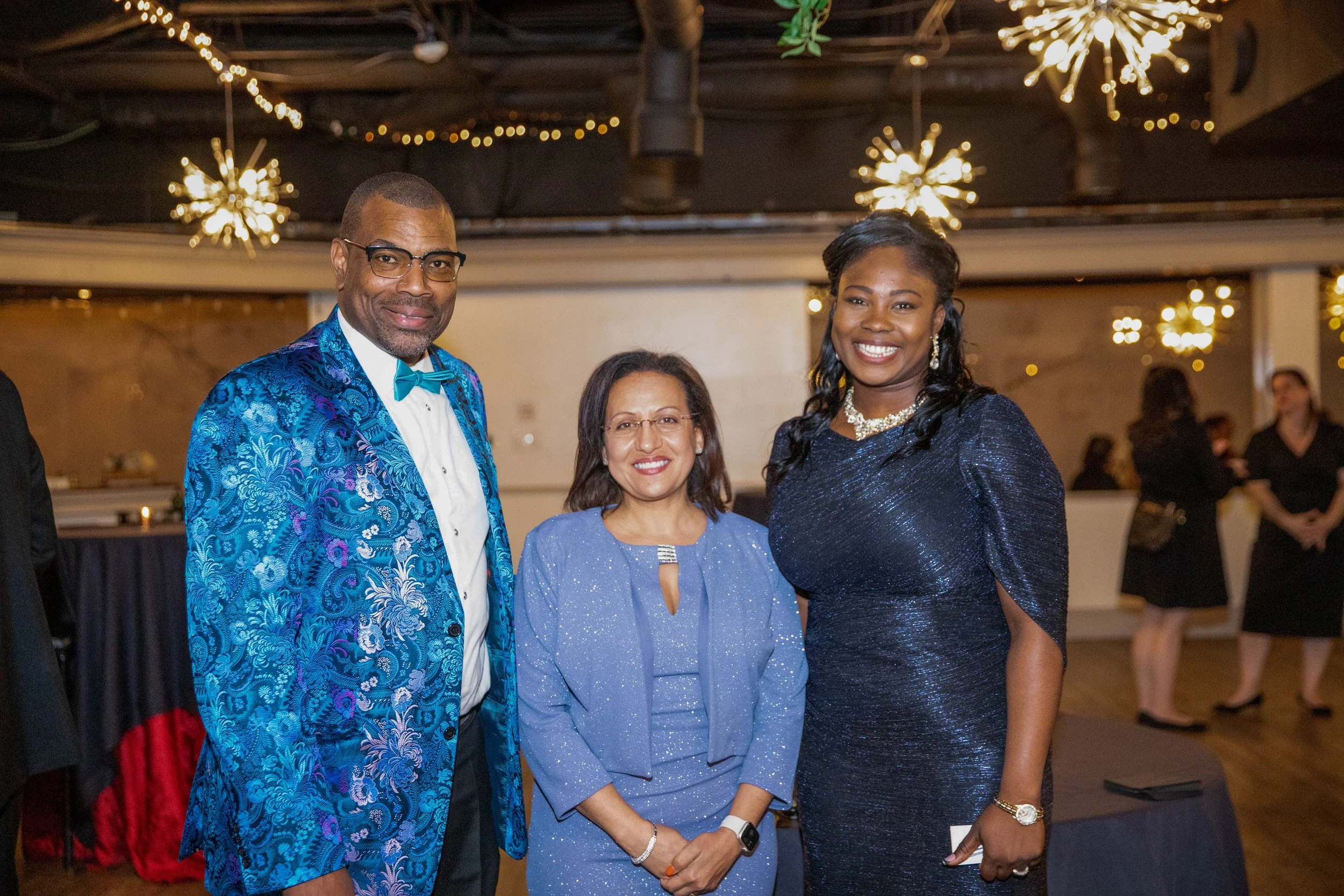 Three women and one man attending a formal event, posing and smiling at the camera. The man is wearing glasses and a vibrant blue patterned jacket with a matching bow tie. One woman is dressed in a sparkling blue dress, another in a dark shimmering d