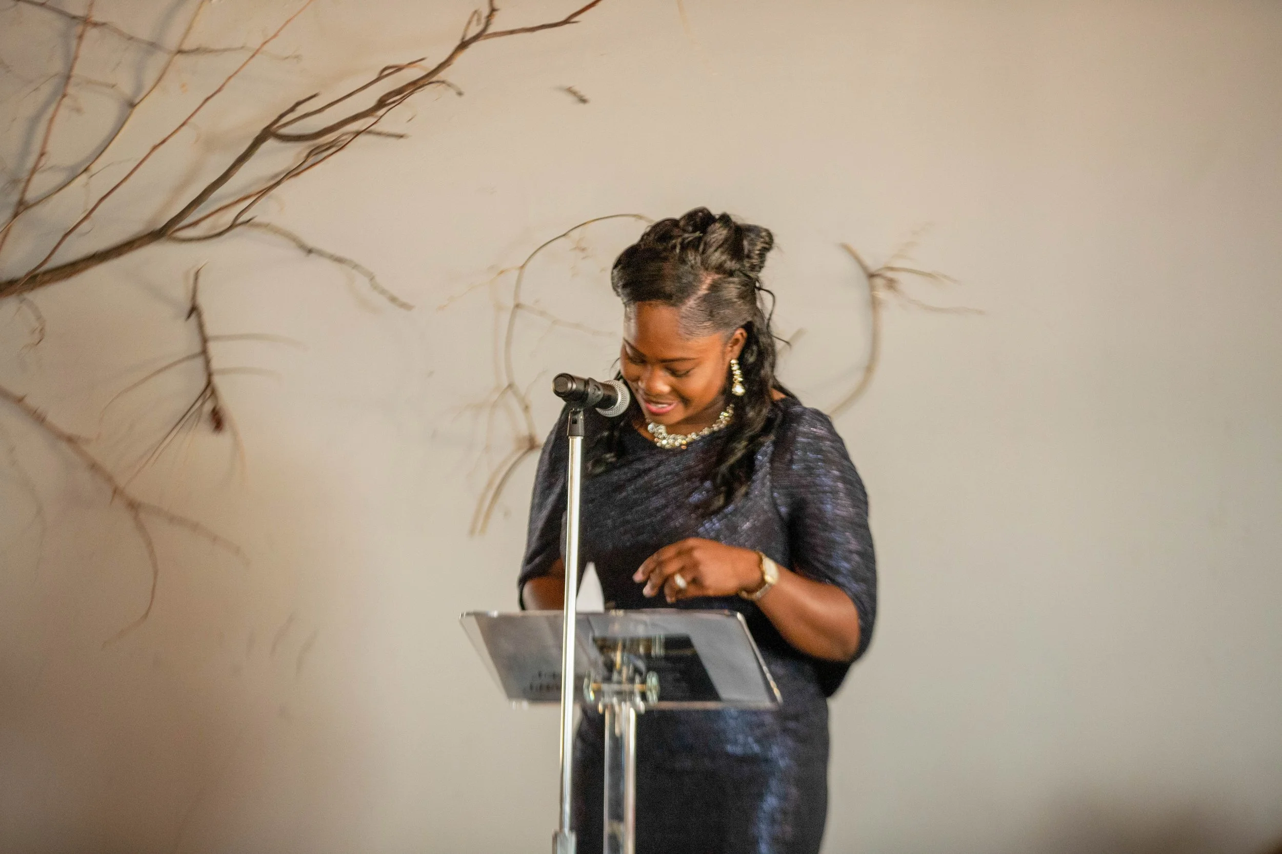 A woman dressed in elegant attire stands behind a lectern, speaking into a microphone, with a neutral-colored wall and decorative branches in the background.