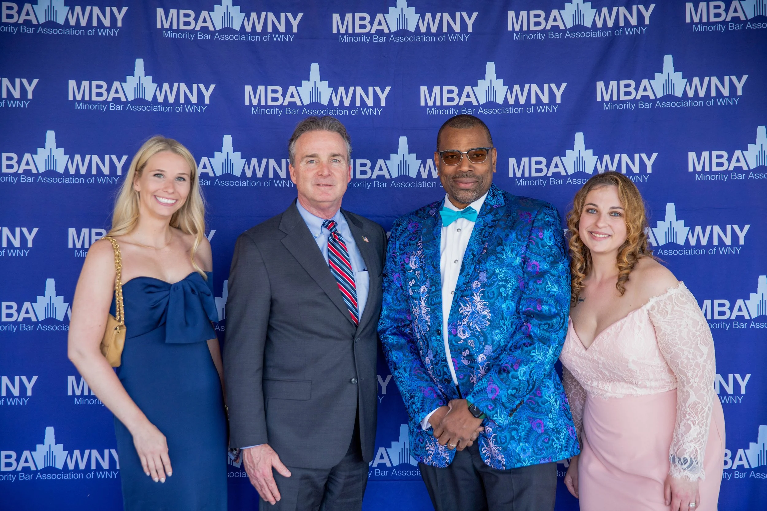 Four people dressed in formal attire standing in front of a blue backdrop with the logos of Minority Bar Association of WNY.