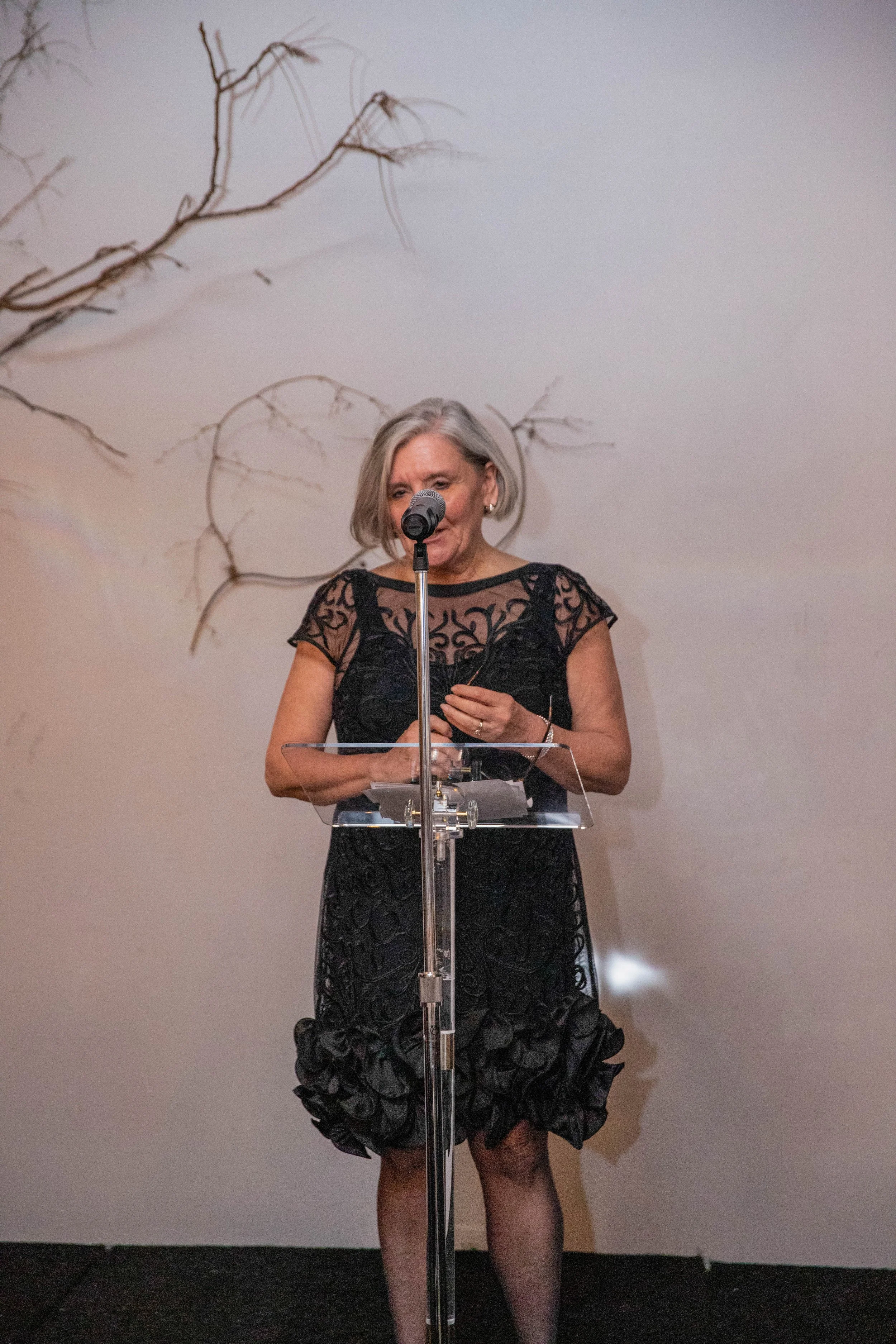 A woman with short grey hair wearing a black lace dress, standing behind a transparent podium, speaking into a microphone, with a plain wall and bare tree branches in the background.