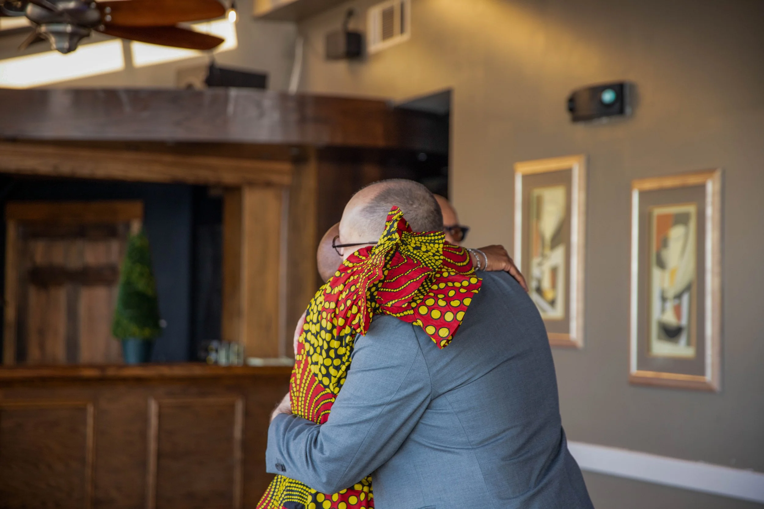 A man in a gray suit is hugging a woman with a colorful patterned scarf in a room with framed pictures and a wooden cabinet.