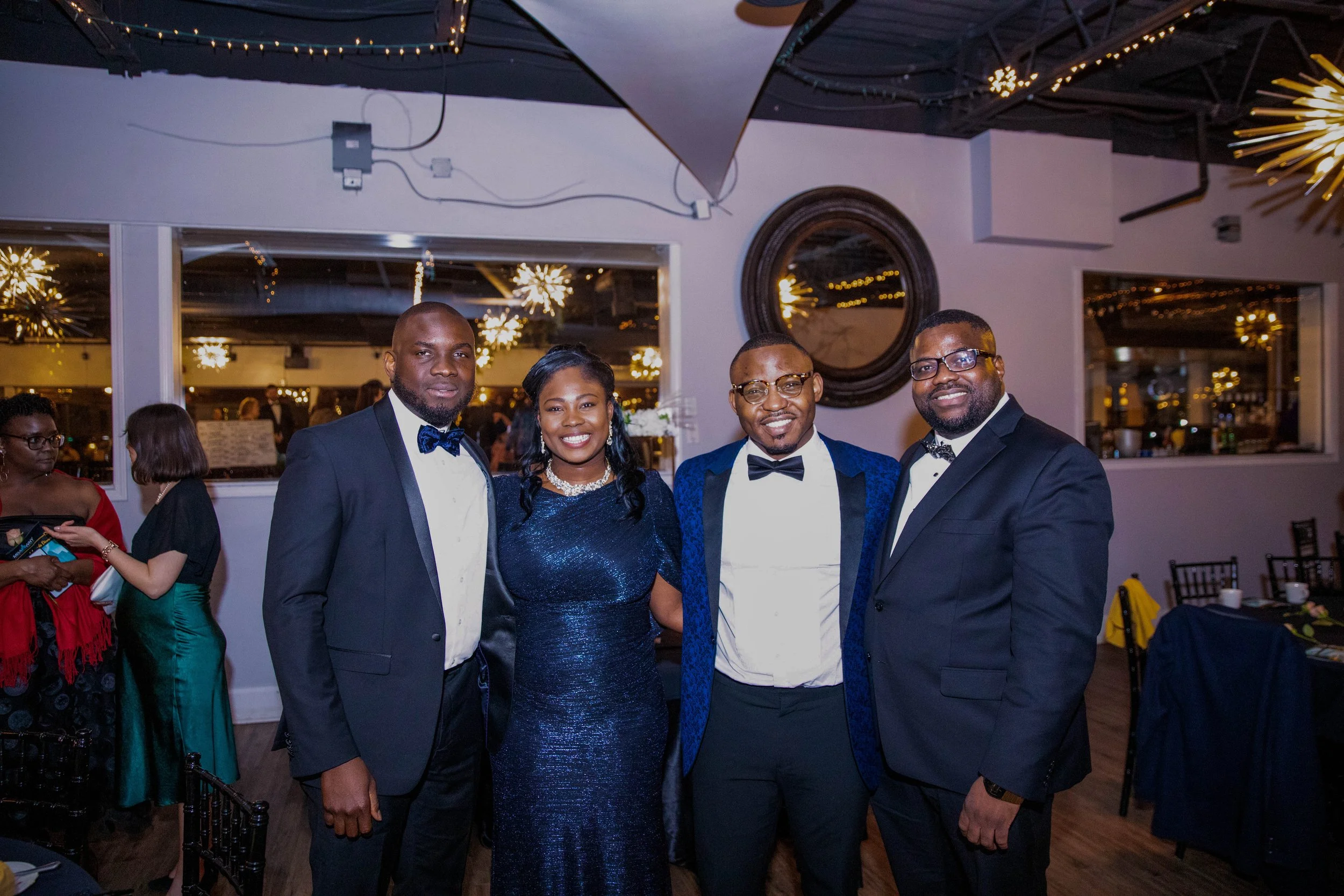 Four well-dressed people, three men and one woman, are smiling at an indoor event, standing closely together. The background shows other guests, large mirrors, and holiday or party lights.