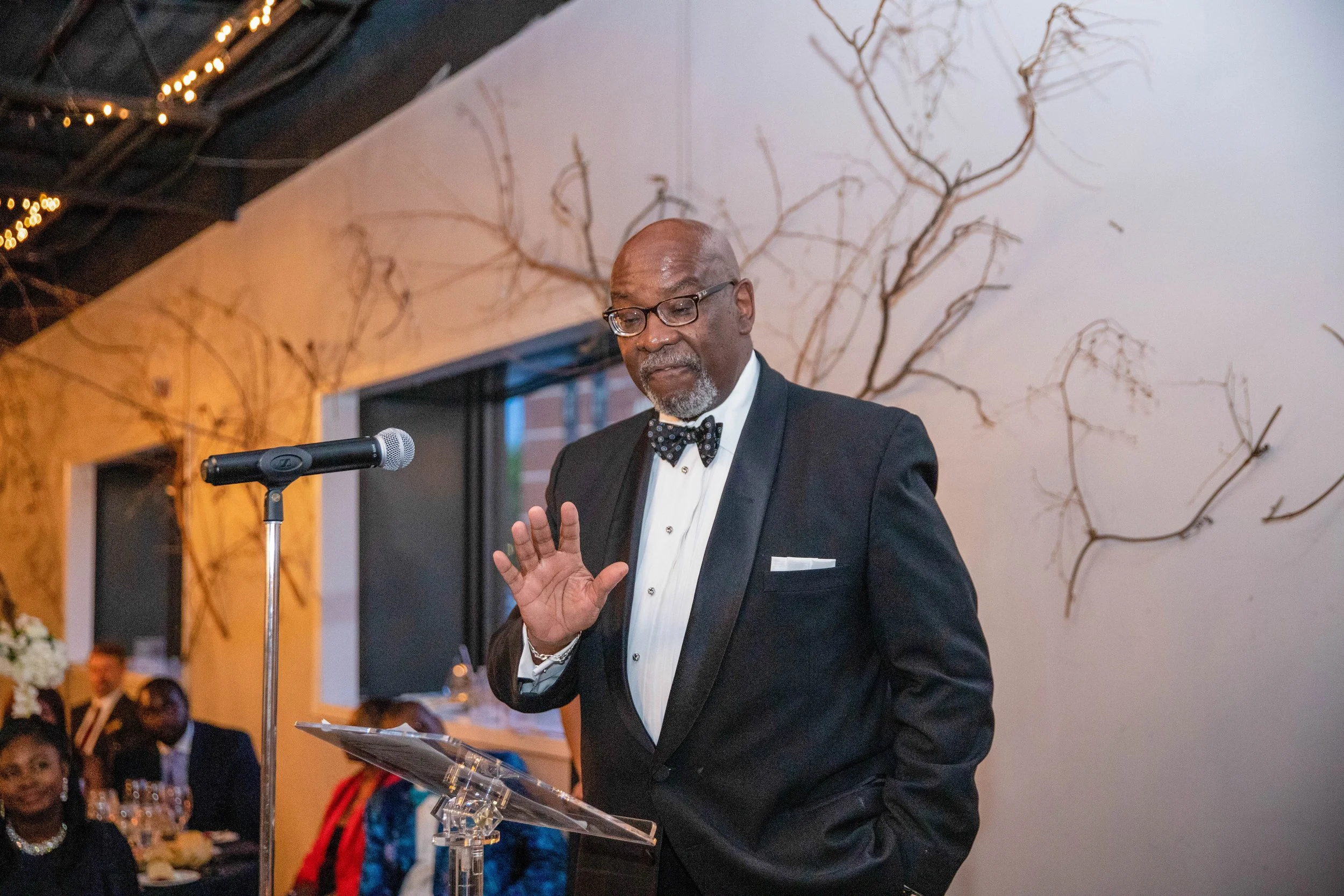 An older man with glasses and a gray beard, dressed in a tuxedo with a bowtie, speaking at a podium during a formal event. He is holding a piece of paper and gesturing with his left hand. The background shows a wall with decorative branches and a cei