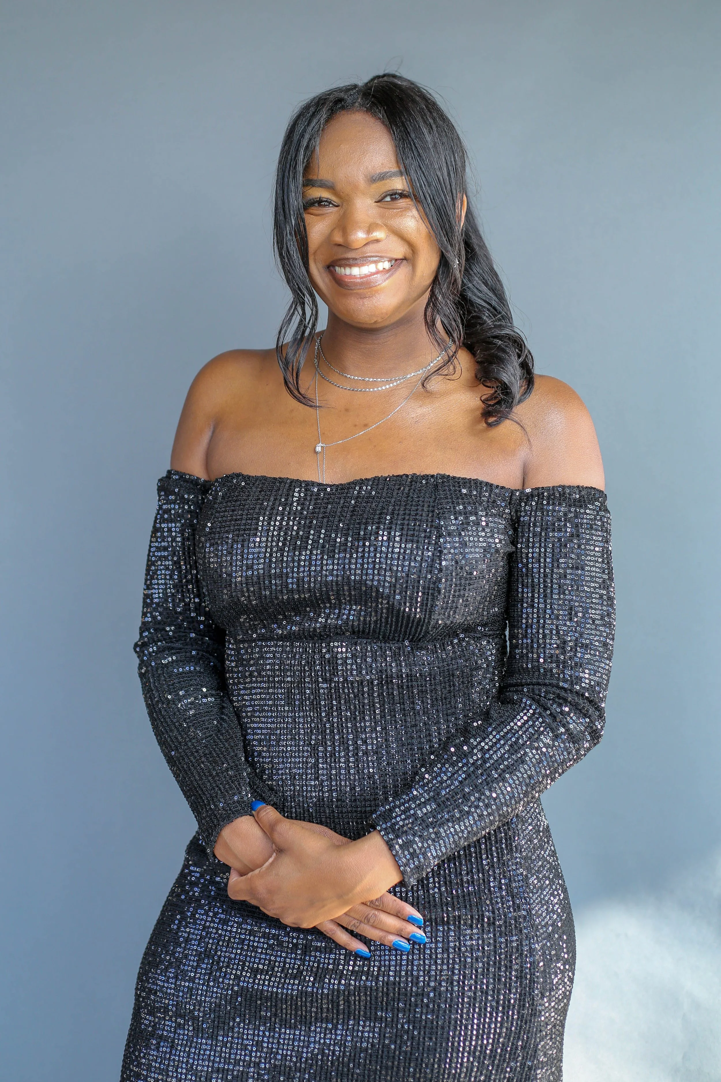 A woman smiling, wearing a black sequined off-shoulder dress, layered silver necklaces, and matching blue and silver nails, standing against a grey background.