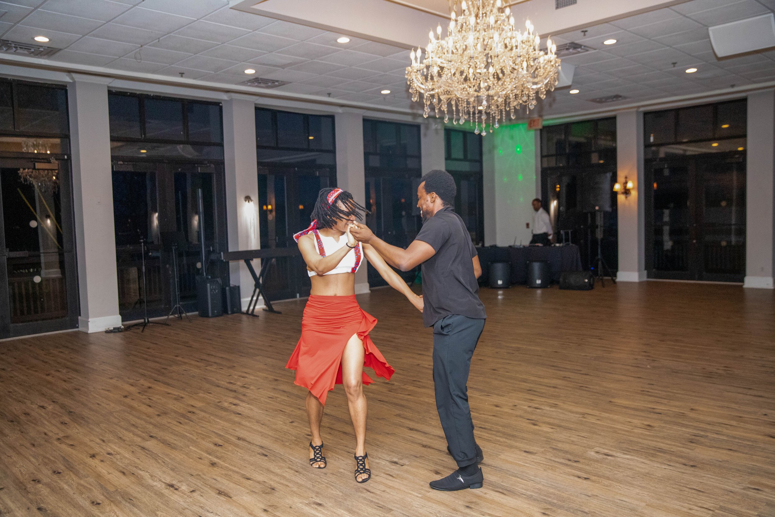 A couple dancing in a ballroom with a large chandelier overhead. The woman is wearing a white top, red skirt, and black heels, while the man is dressed in a black shirt and black pants. The woman is smiling and holding the man's hand, and they appear