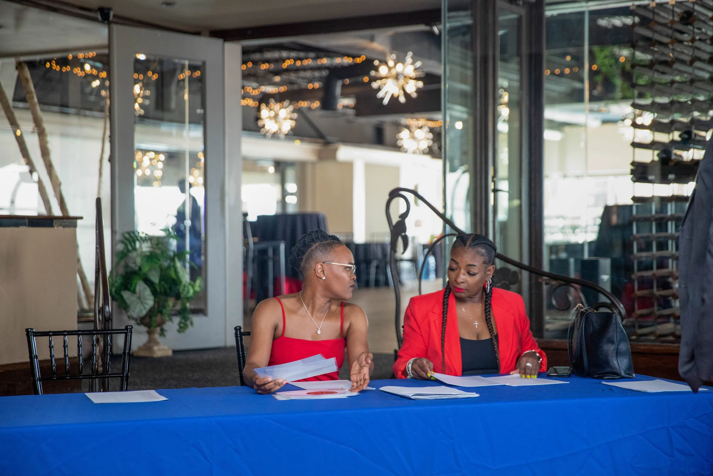 Two women sitting at a table in a modern restaurant or event space, engaged in conversation. The woman on the left has short hair and glasses, wearing a red dress, and holding papers. The woman on the right has braided hair, wearing a red blazer over