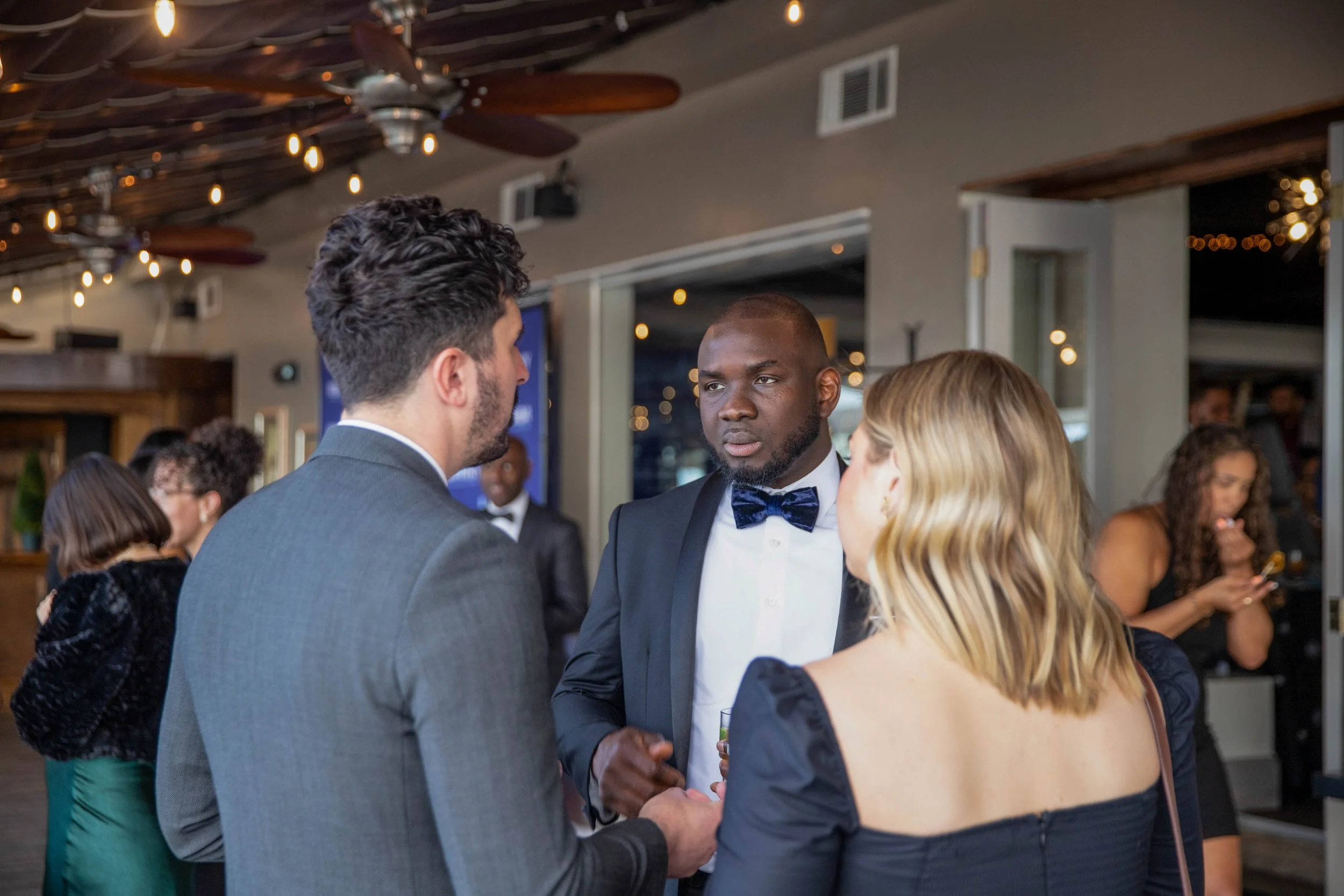 A group of people in formal attire at a social event, including a man in a tuxedo with a blue bow tie, engaged in conversation with others in the background.