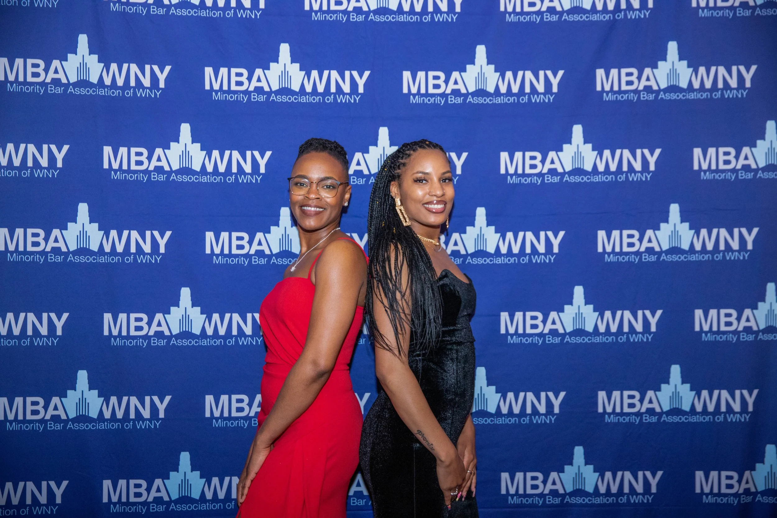 Two women in elegant dresses standing in front of a blue backdrop with white text and logos for the Minority Bar Association of WNY. One woman is wearing a red dress and glasses, the other a black dress with earrings.