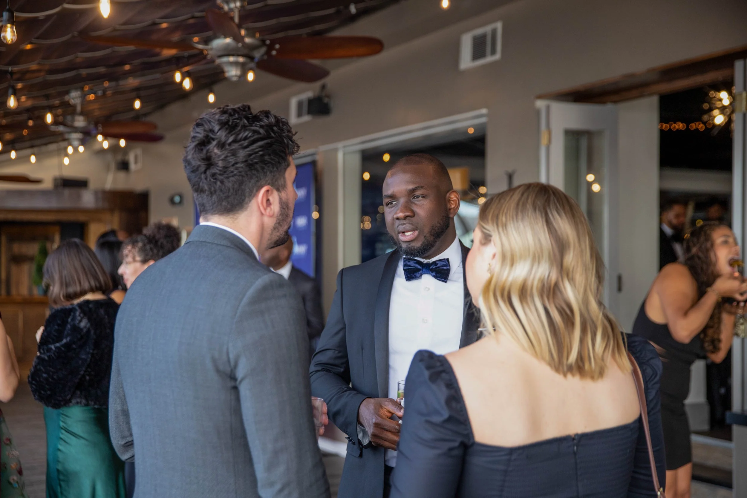 People at a formal event socializing indoors, with lighting and ceiling fans visible.