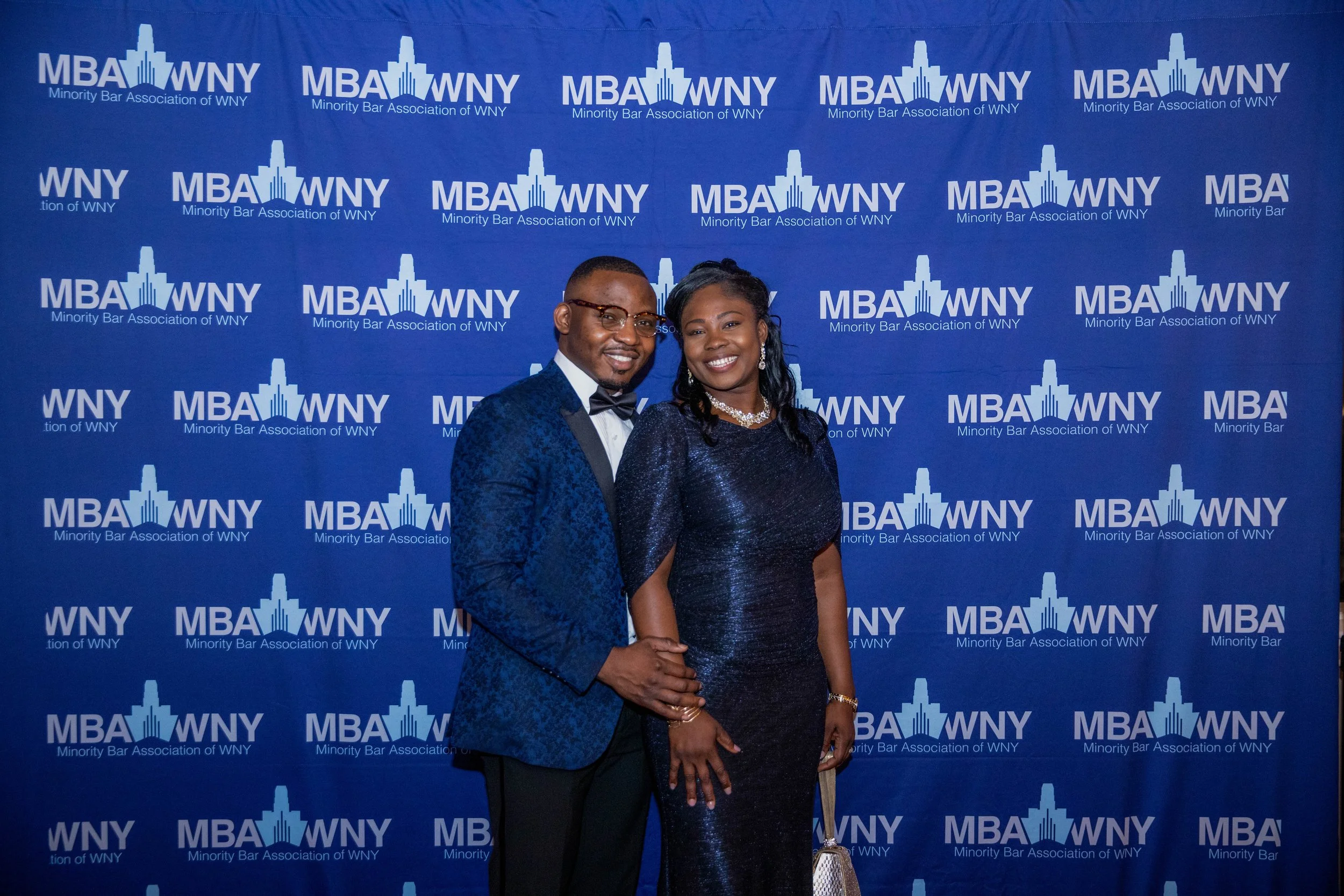 A man and woman in formal attire standing in front of a blue backdrop with white text and logos that read 'MBA WNY Minority Bar Association of WNY.' The man is wearing glasses, a tuxedo with a bow tie, and the woman is wearing a dark, sparkly gown wi