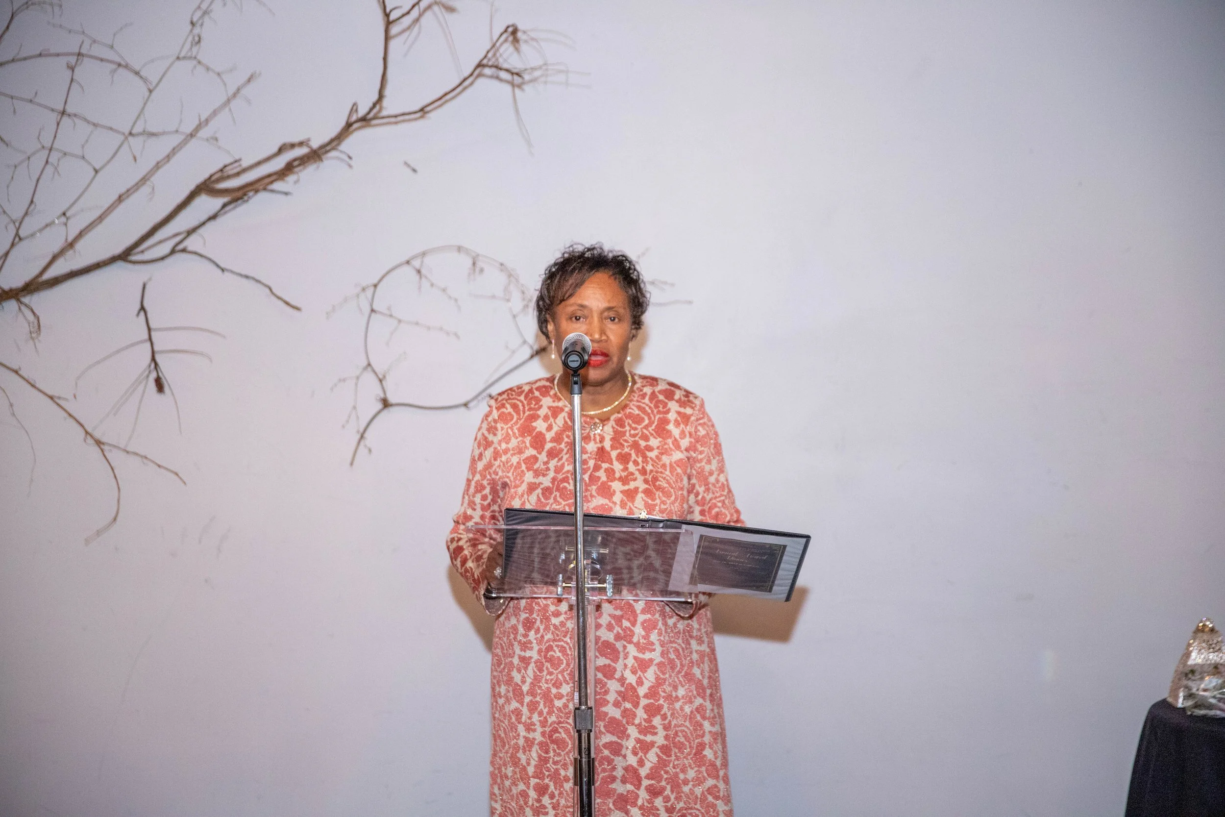 A woman in a coral patterned dress stands behind a clear podium, speaking into a microphone, with a white wall and bare tree branches in the background.