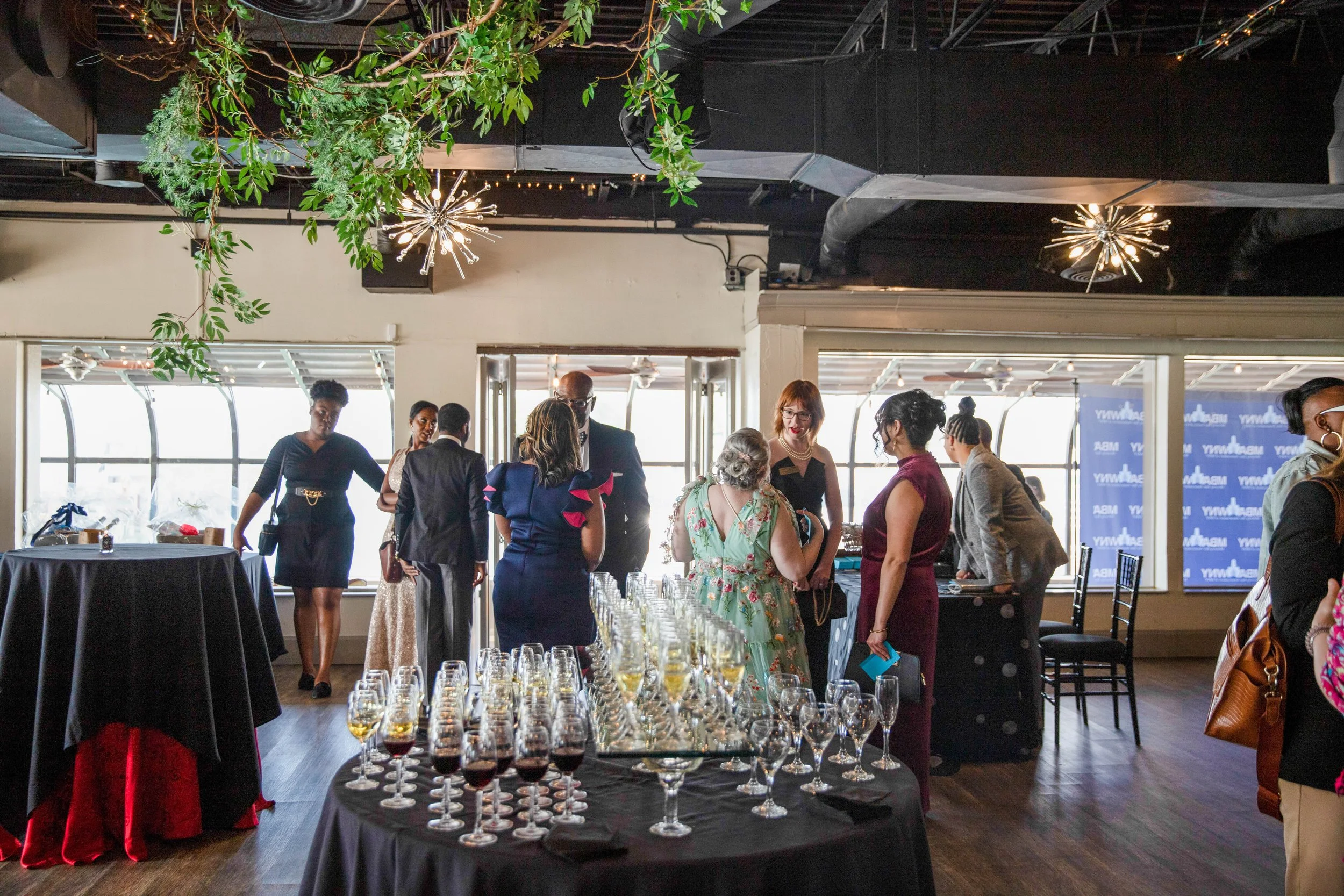 Indoor event space with a group of people socializing, a table with numerous glasses of wine, and decorative hanging lights. Large windows in the background show a view outside.