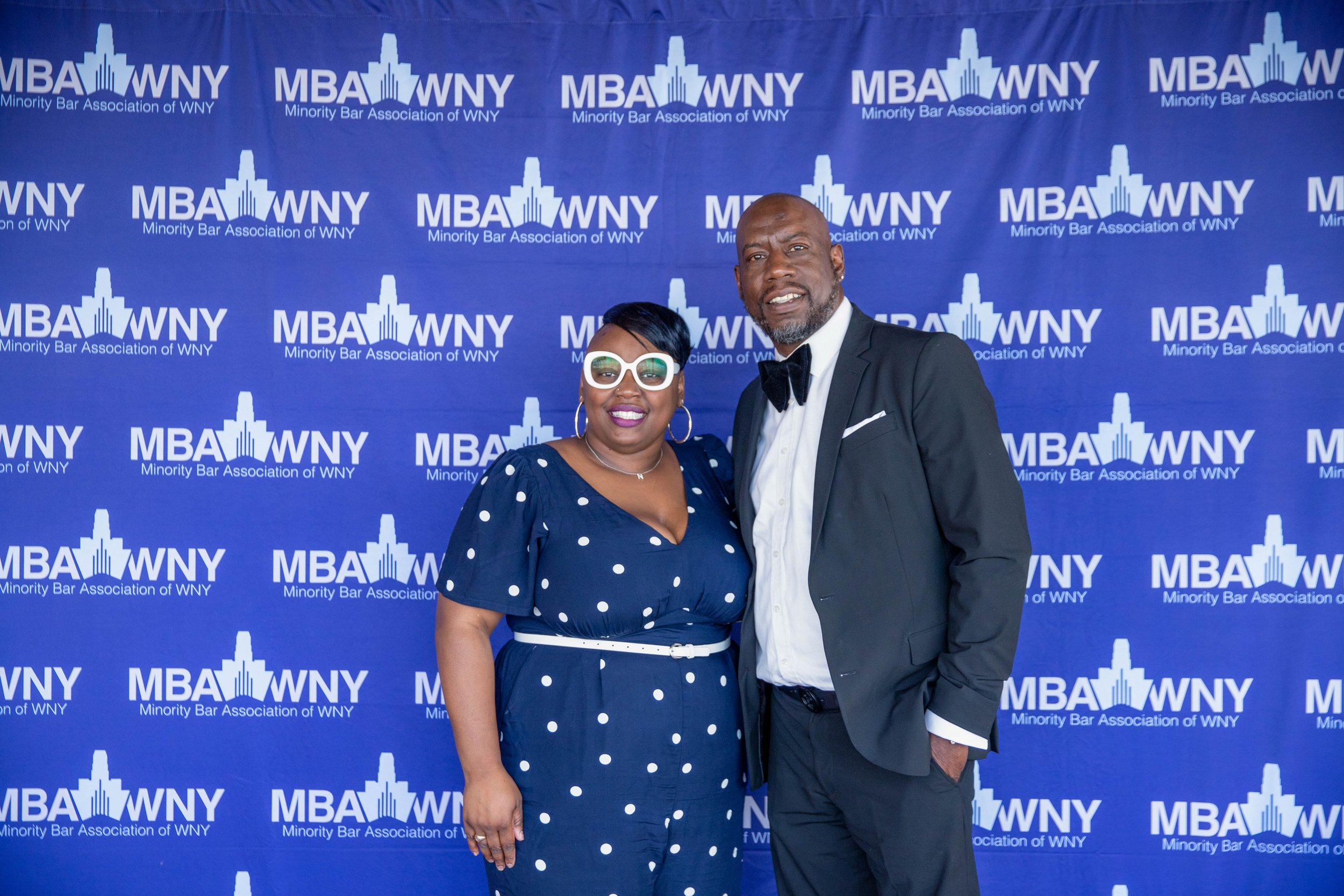 A woman and a man dressed in formal attire standing in front of a blue backdrop with the logo and text 'MBA WNY Minority Bar Association of WNY'. The woman is wearing a navy blue polka dot dress with a white belt, large glasses, and gold hoop earring