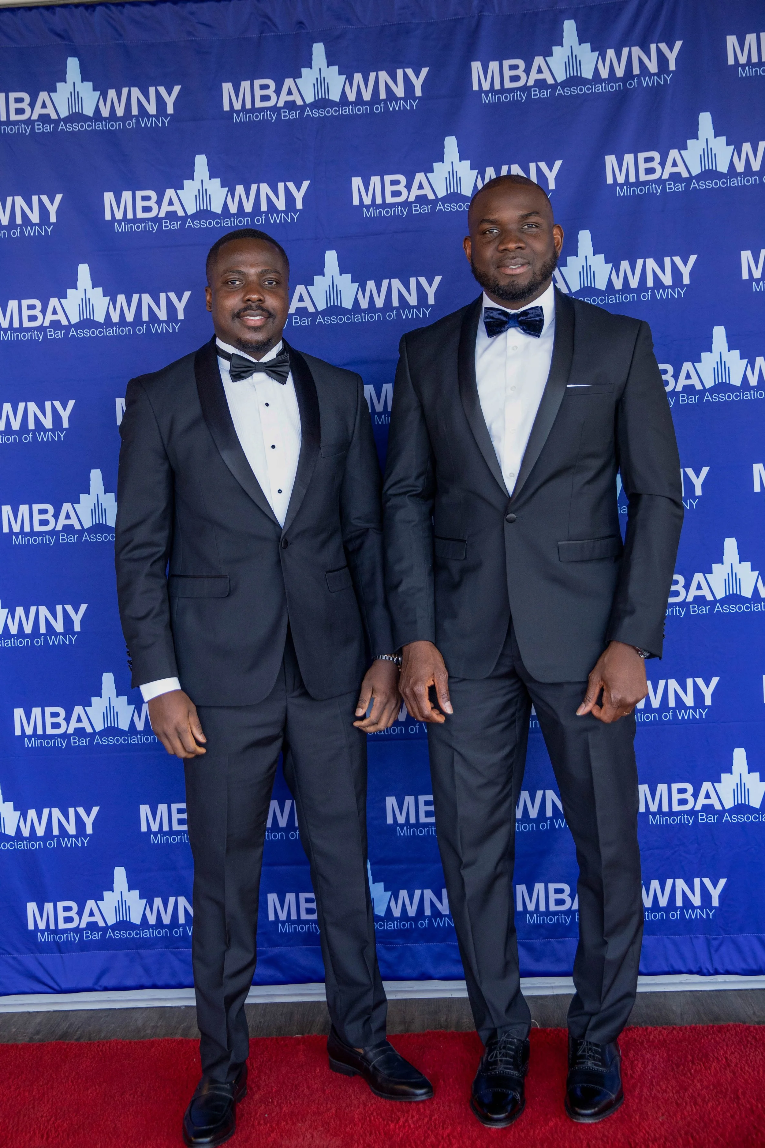 Two men in tuxedos standing on a red carpet in front of a blue backdrop with the text "MBA WNY, Minority Bar Association of WNY."