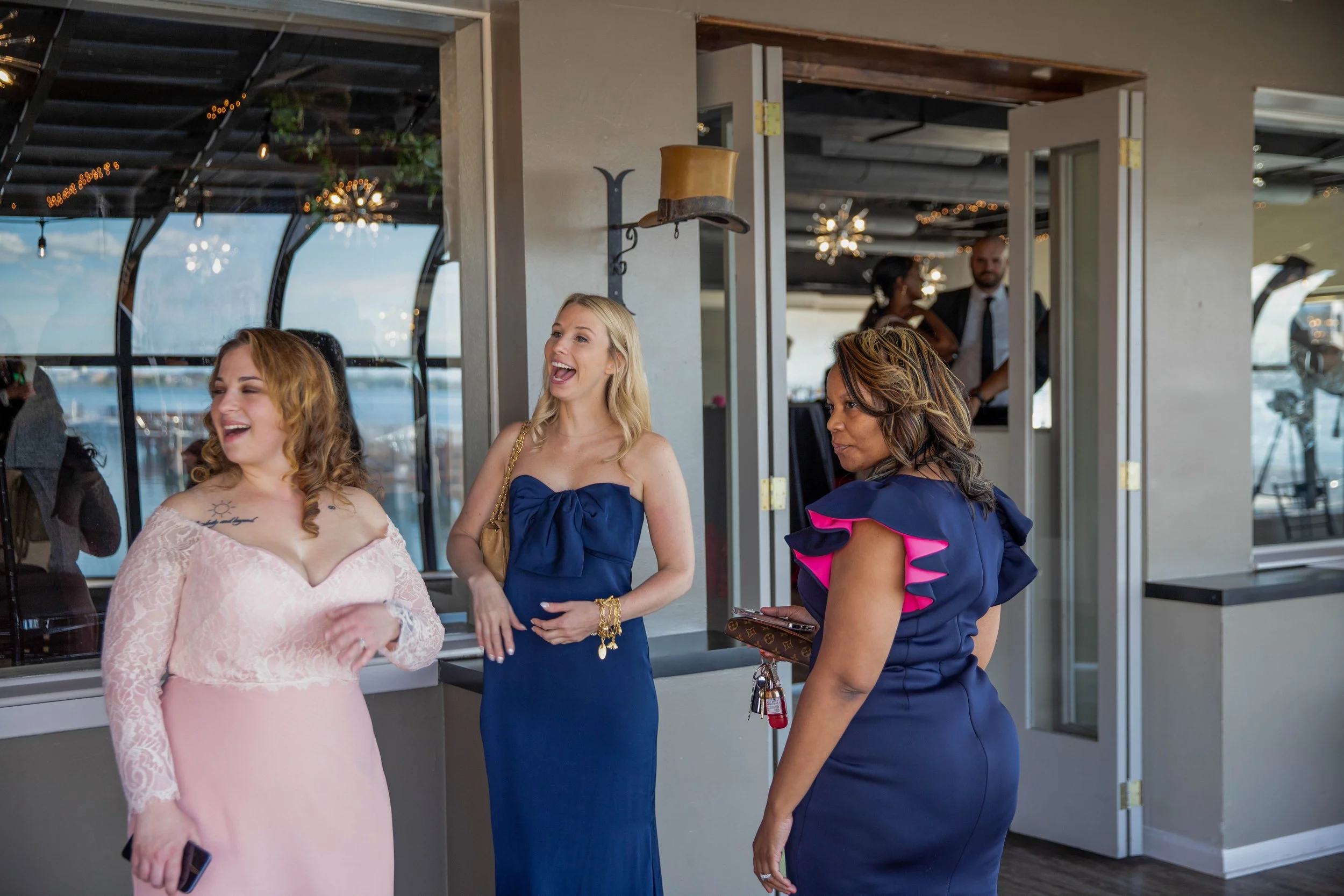 Three women are standing and talking inside a modern building with large windows. They are dressed in formal attire, smiling, and appear to be enjoying a social event or gathering. Two of the women are facing each other, while the third woman, on the