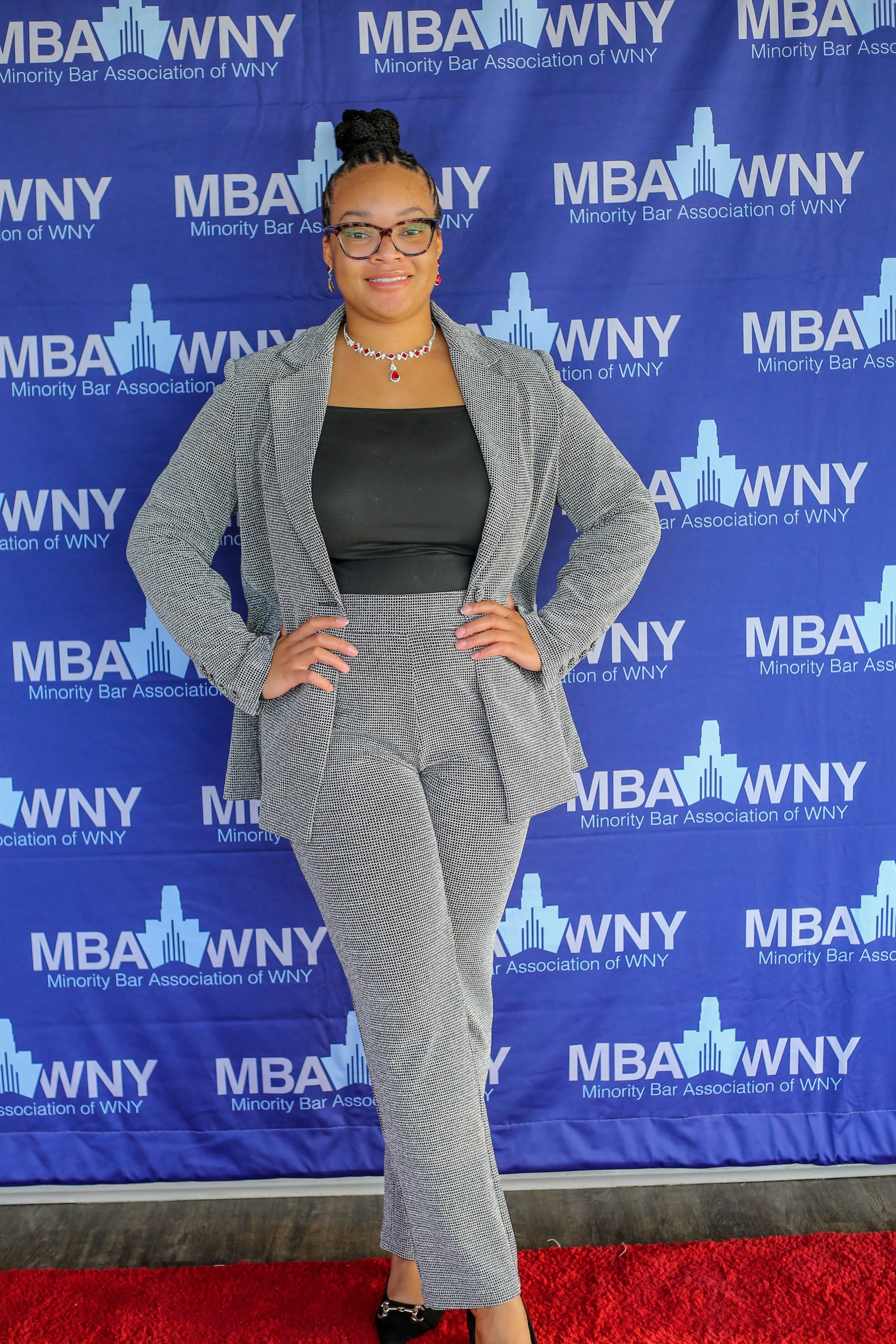 A professional woman standing confidently with hands on hips in front of a blue backdrop with the logos of MBA WNY, the Minority Bar Association of WNY. She is wearing glasses, a grey plaid blazer and pants, a black top, and jewelry, including a neck