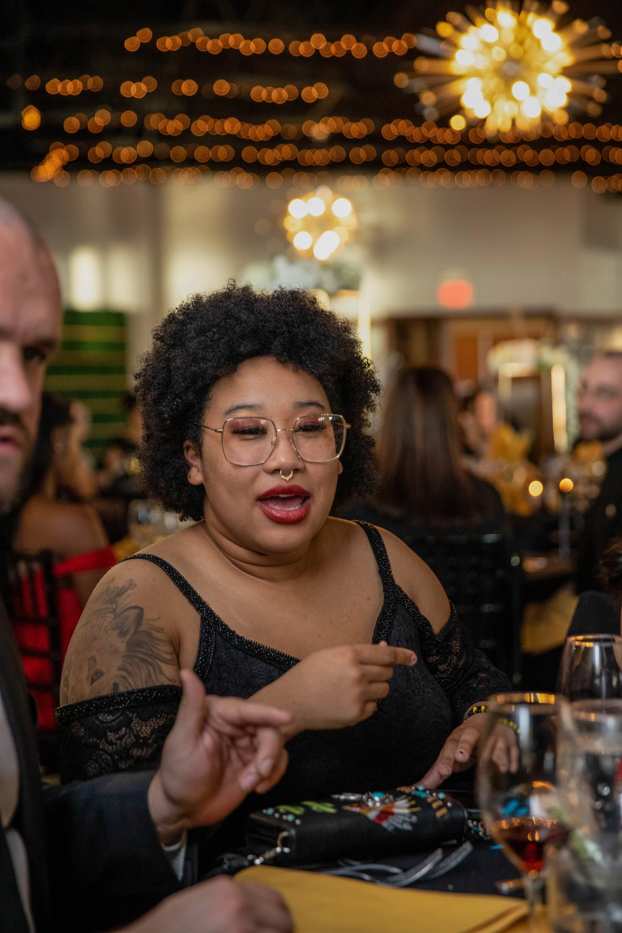 A woman with glasses and curly hair talking at a social gathering indoors with warm lighting and hanging decorations.
