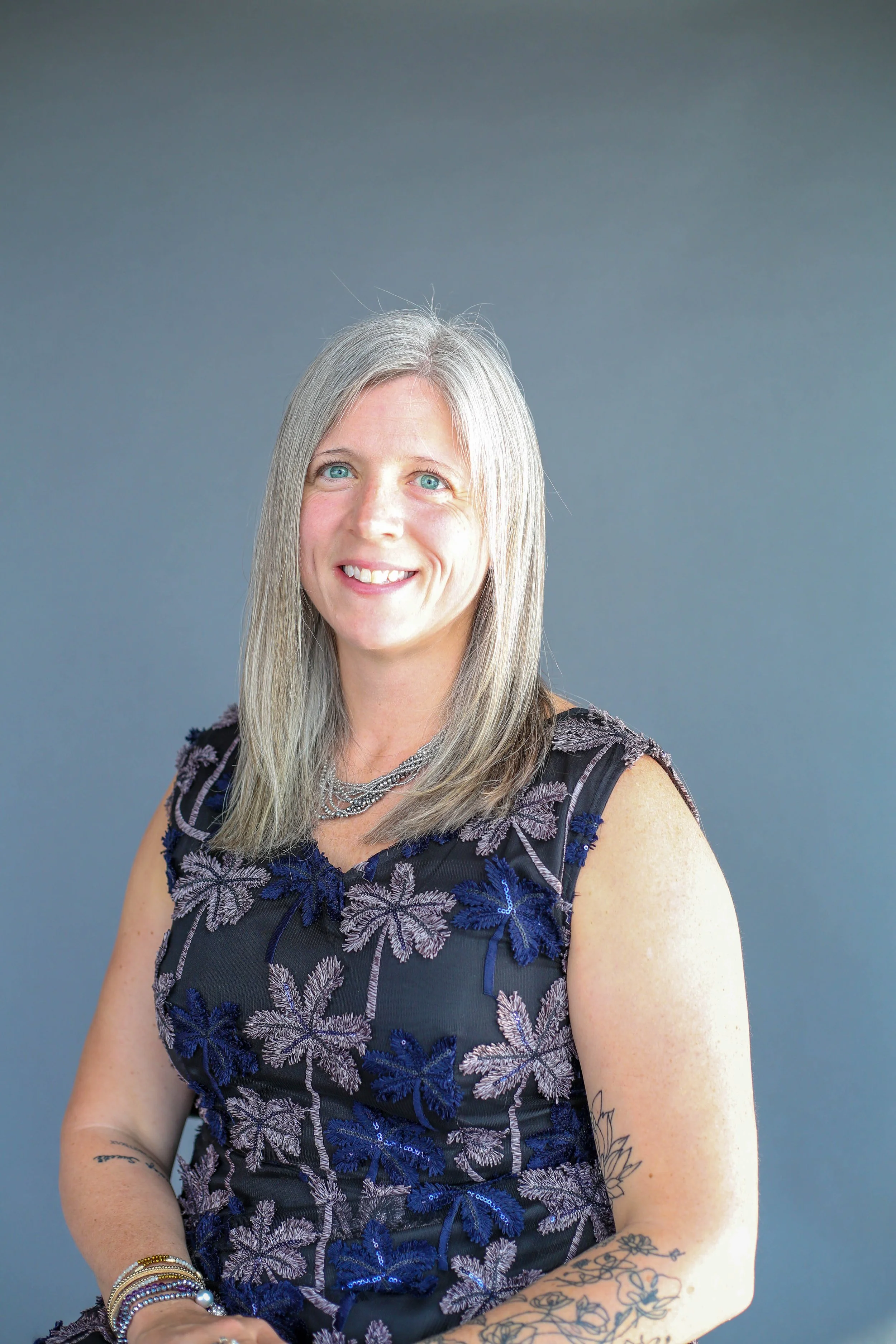 Portrait of a woman with long gray hair, blue eyes, wearing a black dress with purple and blue floral embroidery, and multiple bracelets, sitting against a gray background.