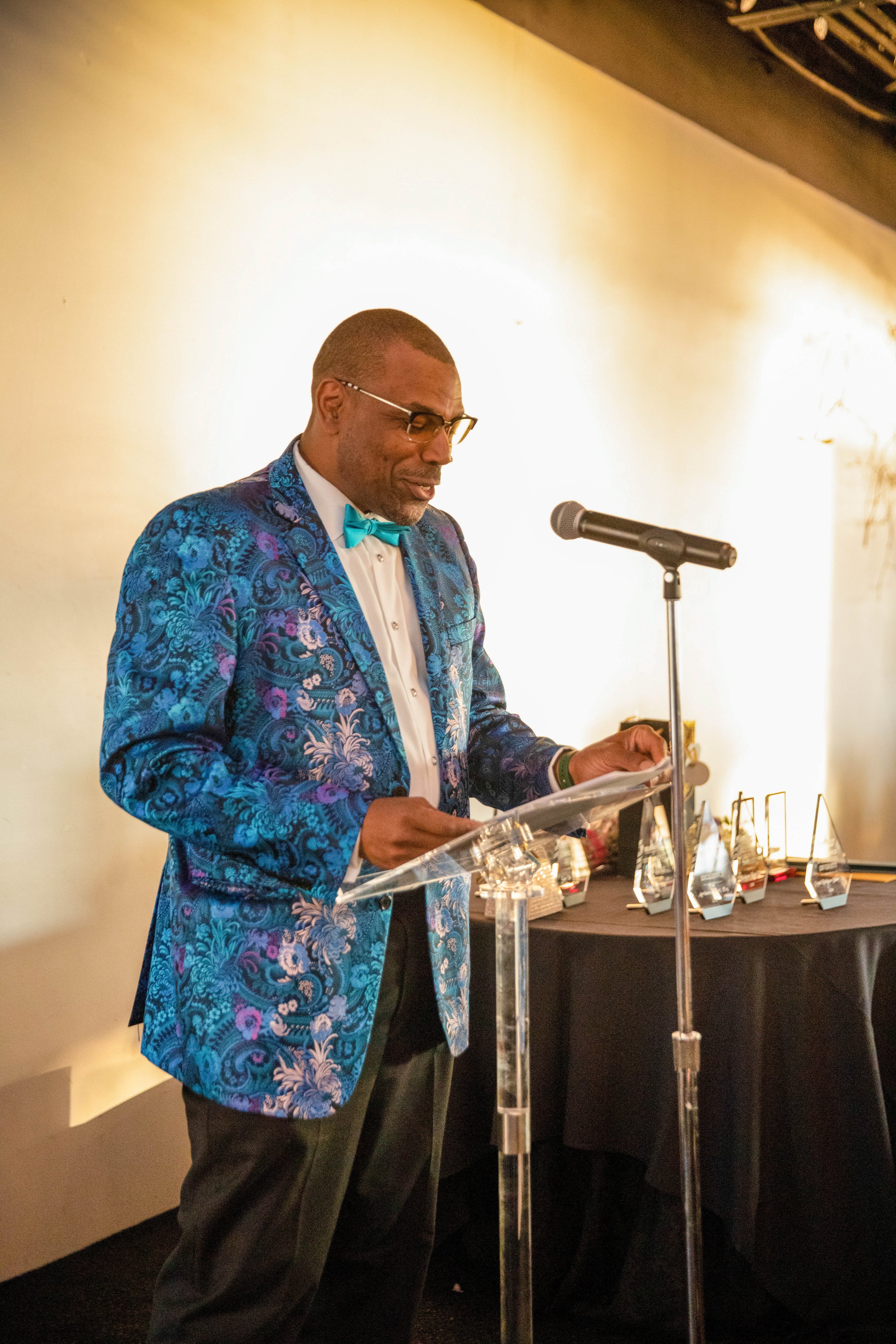 A man in a blue floral blazer, white shirt, and turquoise bow tie is standing at a podium and reading from paper in front of a microphone. There are awards or trophies displayed on a table behind him.