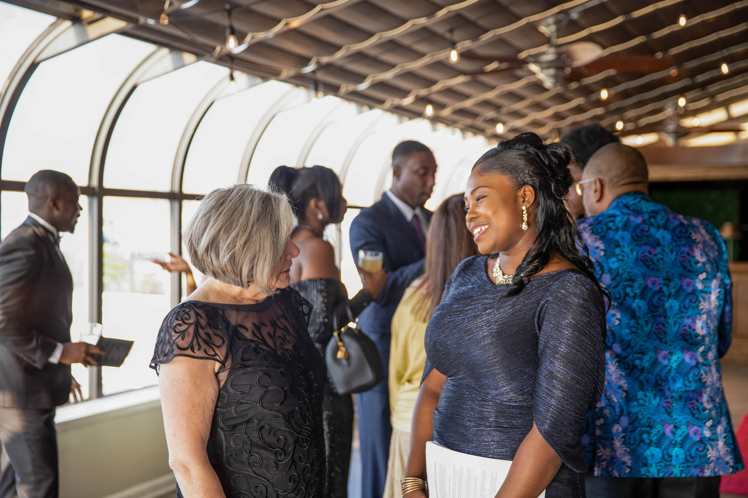 People socializing at a formal event, some holding papers or drinks, in an indoor venue with large windows and string lights overhead.
