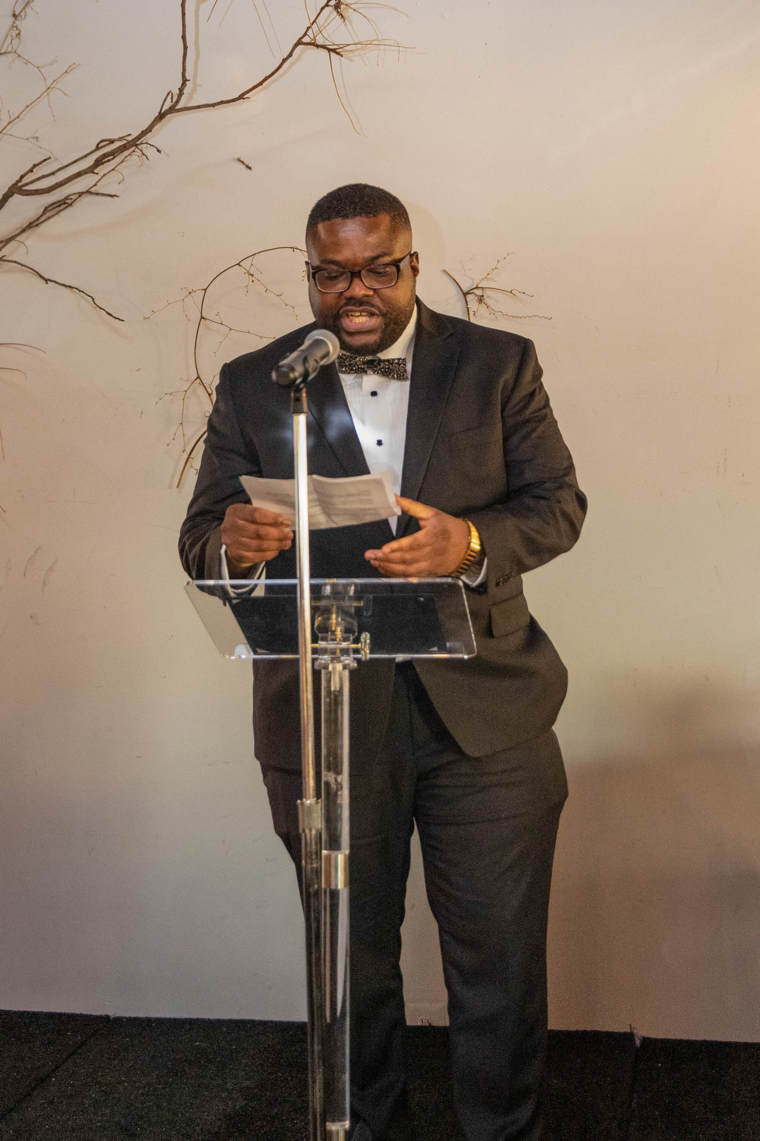 A man wearing a black tuxedo, white shirt, and bow tie stands at a clear podium, reading from a paper with a microphone in front of him. He has short hair, glasses, and a gold watch. Behind him, there are decorative, dry, leafless branches on a plain