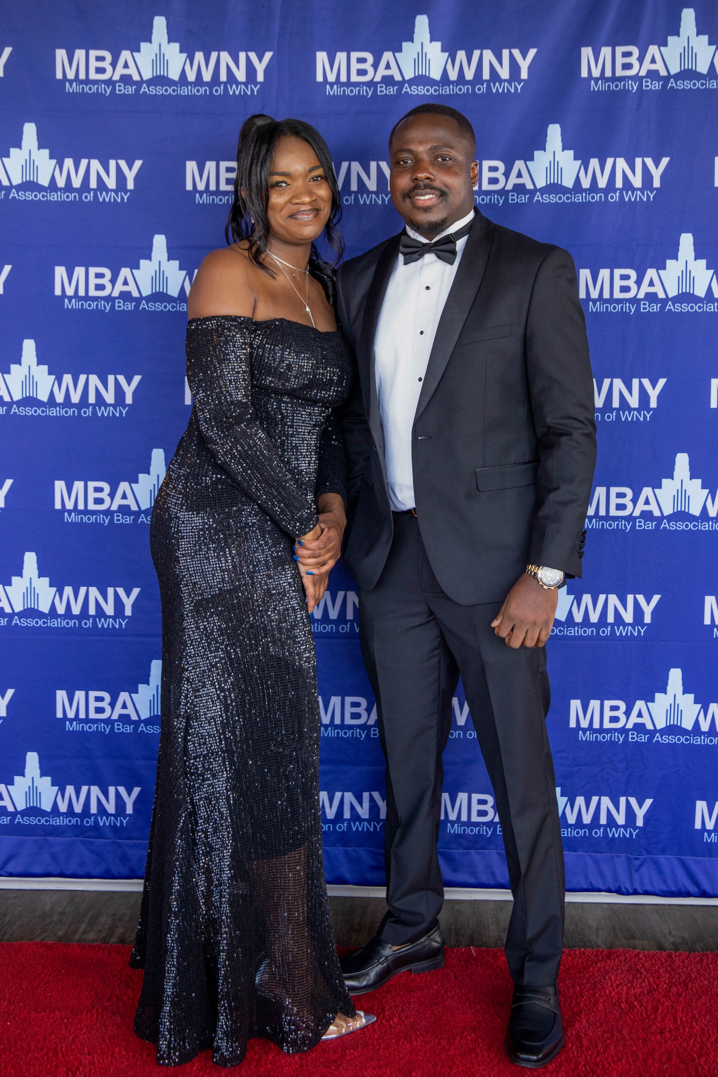 A man and woman dressed in formal attire standing on a red carpet in front of a blue backdrop with the logo of the Minority Bar Association of WNY.