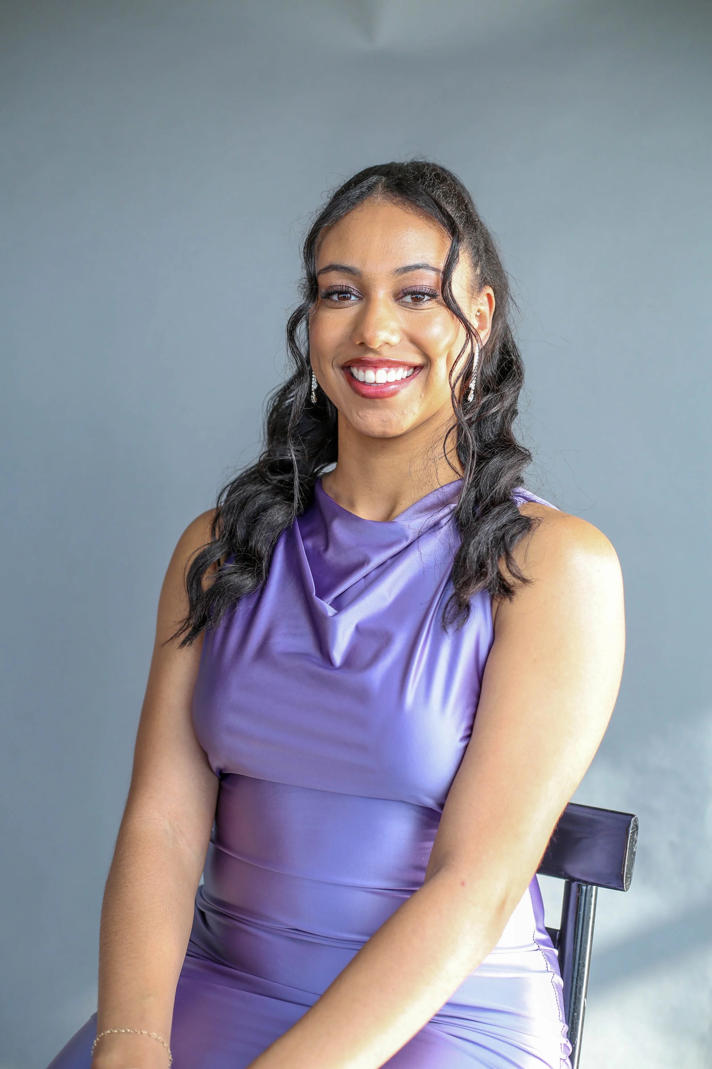 A woman smiling, wearing a purple satin dress, seated on a black chair against a gray background.