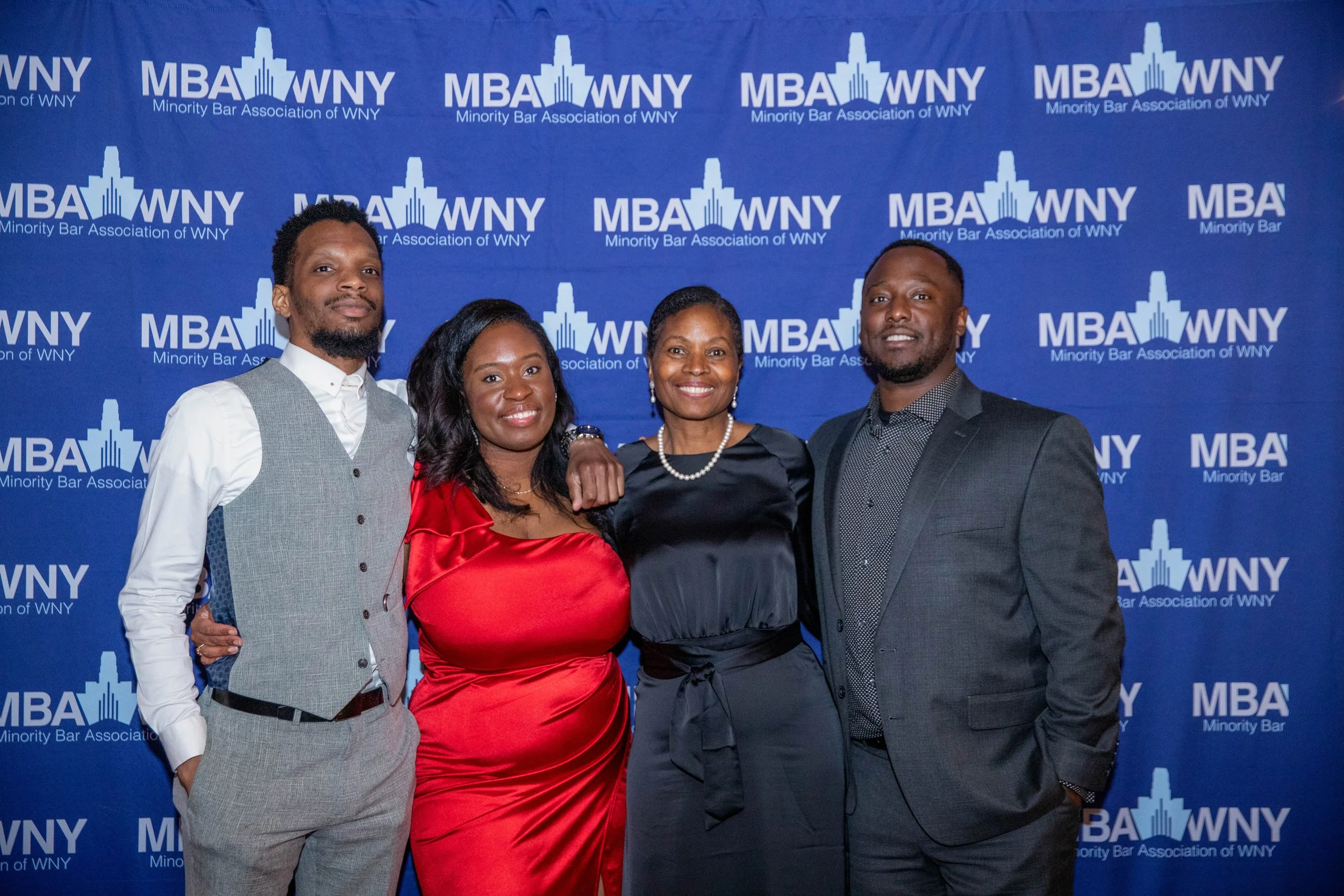 Four people standing in front of a blue backdrop with the logo and text 'Minority Bar Association of WNY.' They are dressed formally and smiling at the camera.
