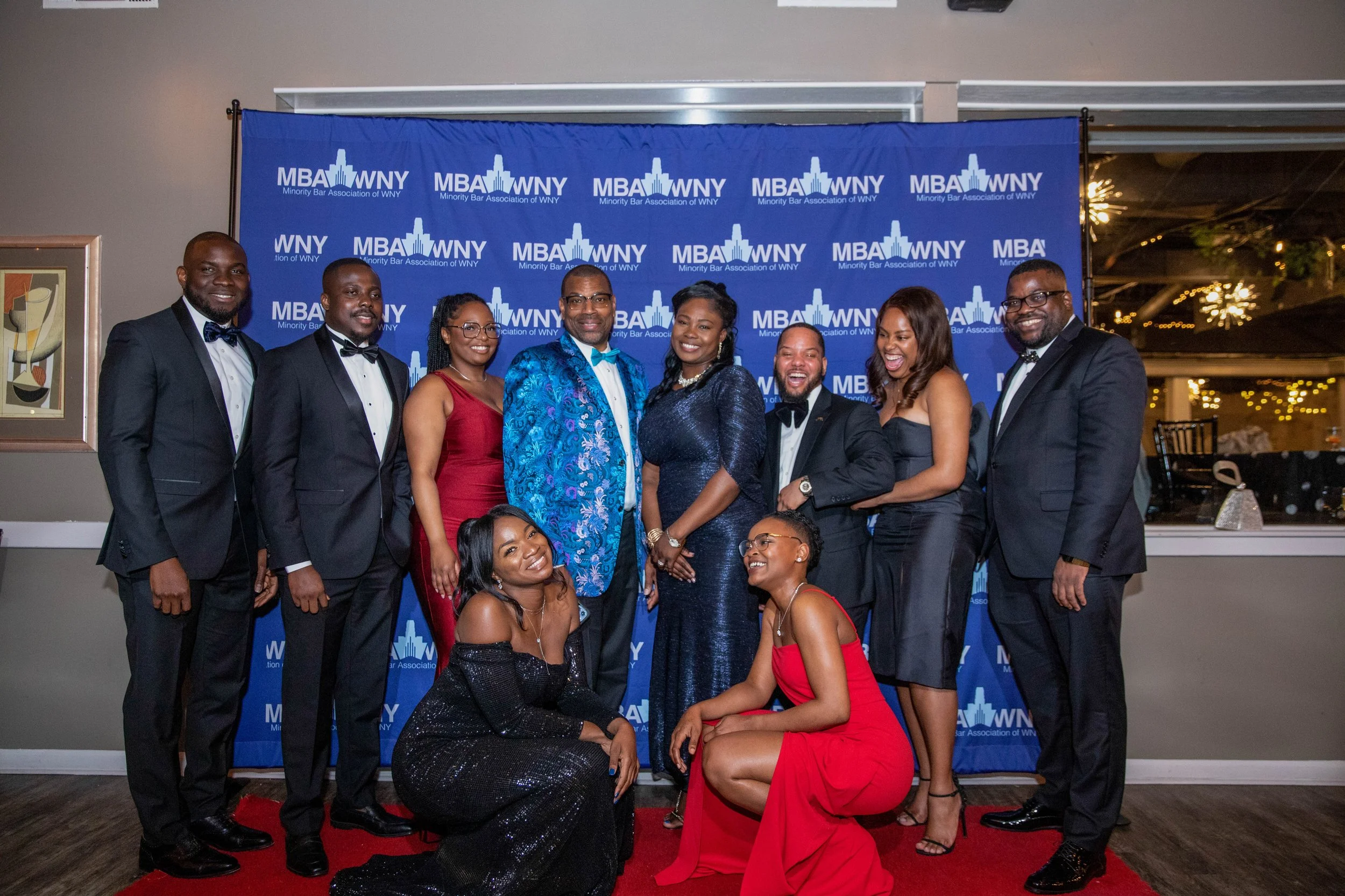 Group of nine well-dressed people at a formal event, smiling and posing in front of a blue backdrop with the logo of the Minority Bar Association of WNY, indoors with warm lighting and festive decorations.
