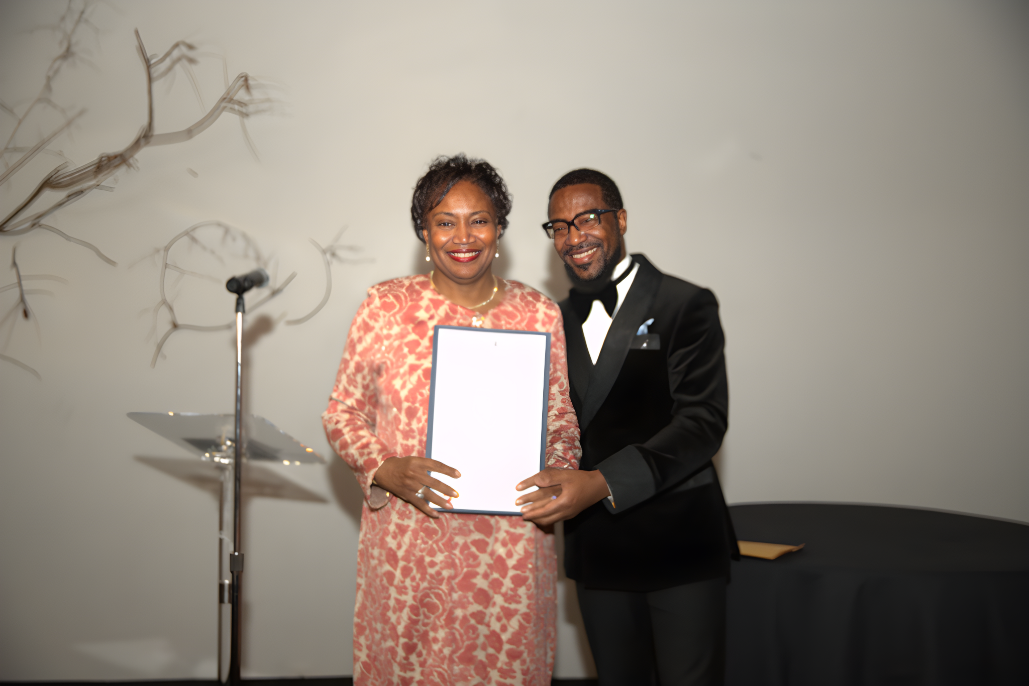A woman in a pink dress holding a certificate, standing next to a man in a black tuxedo with glasses, both smiling at a formal event.