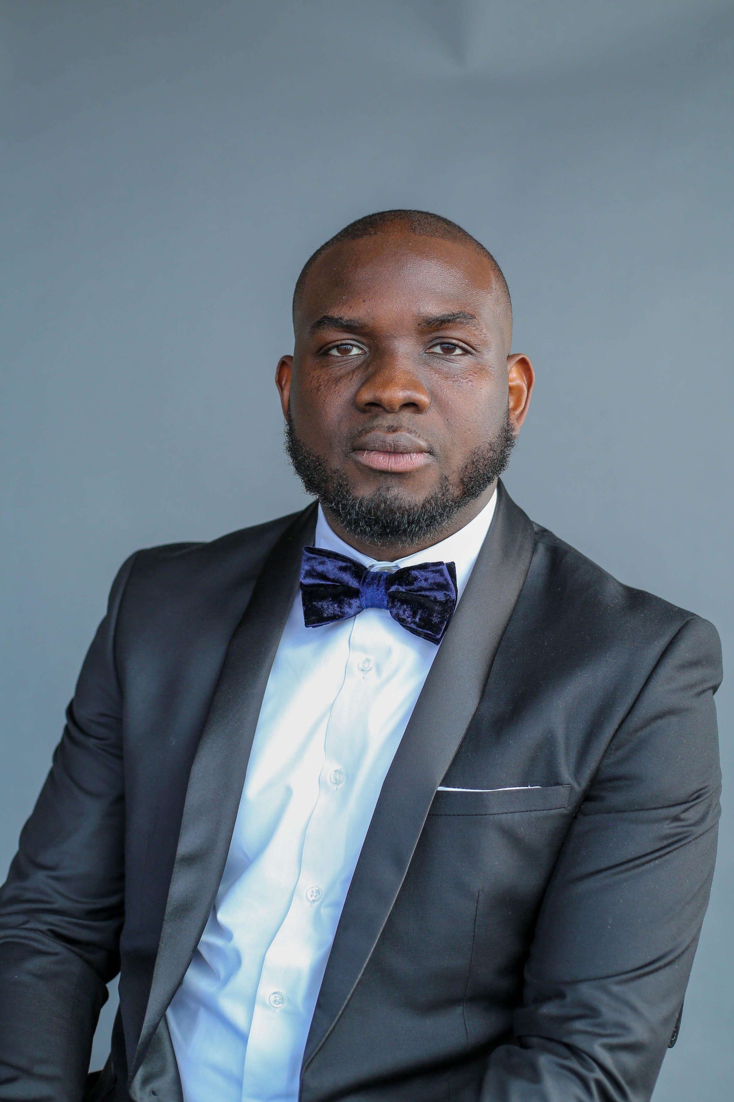 A man in a black tuxedo, white shirt, and purple velvet bow tie posing against a plain grey background.