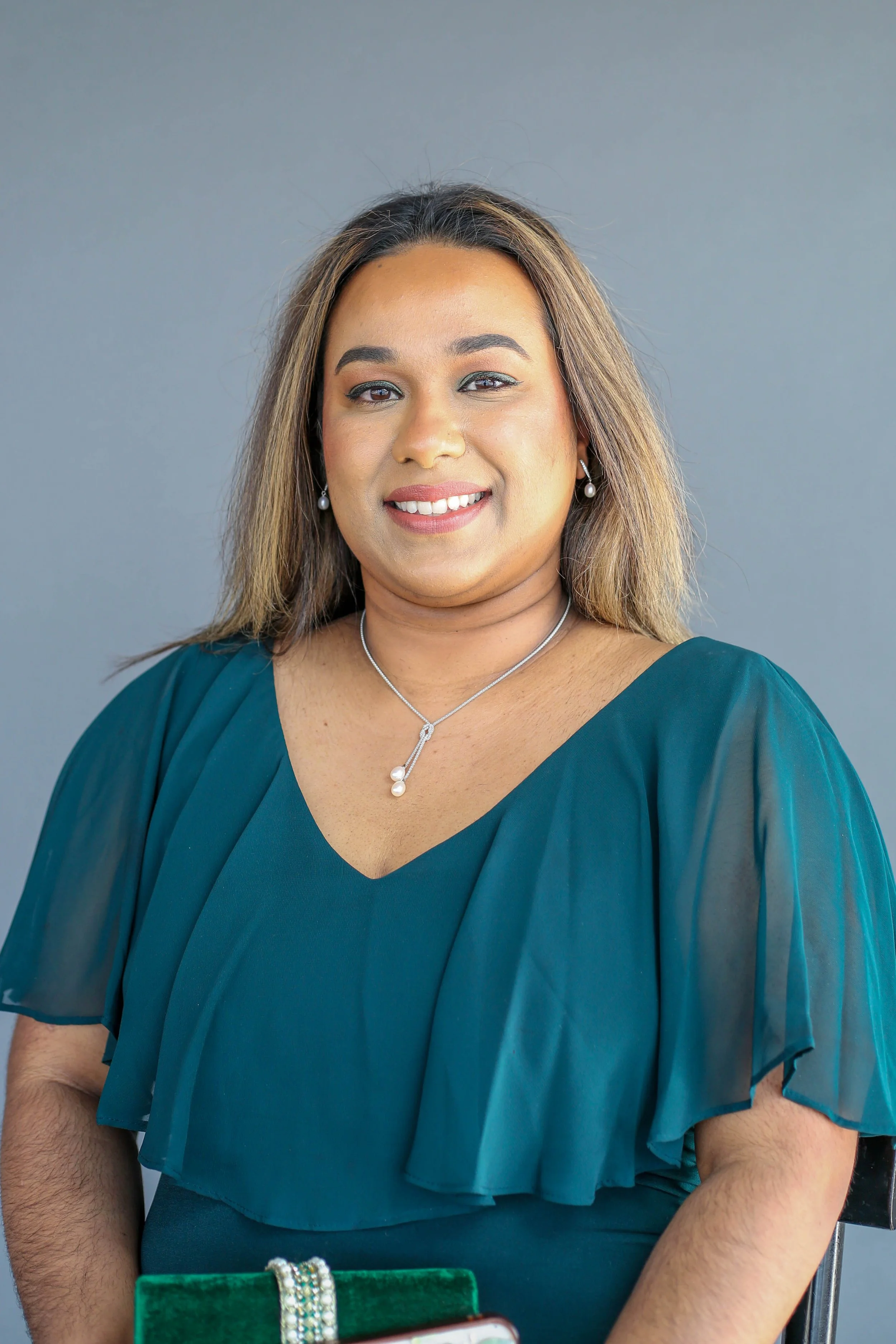 A woman with long, light brown hair smiling, wearing pearl earrings, a silver necklace, and a teal dress with layered ruffled sleeves, standing in front of a plain gray background.