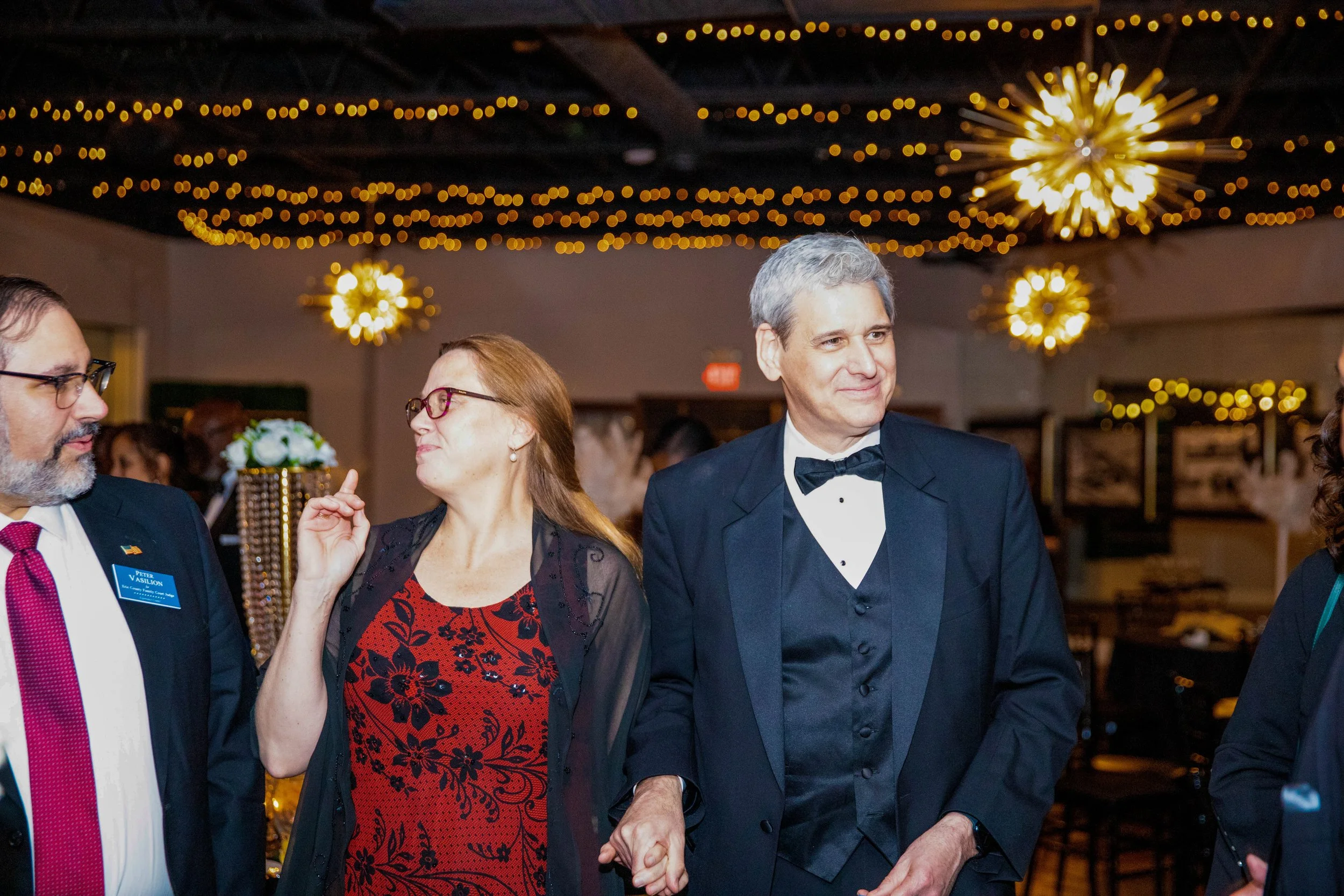 People dressed in formal attire socializing at a festive event with string lights and decorative chandeliers overhead.
