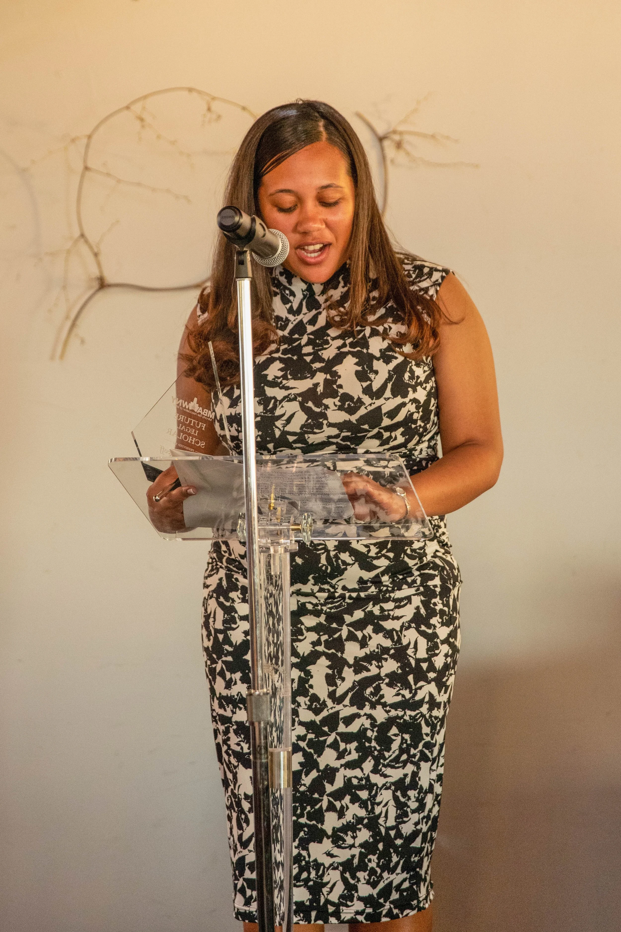 Woman in a black and white patterned dress standing at a podium, reading or speaking into a microphone against a light-colored wall background.