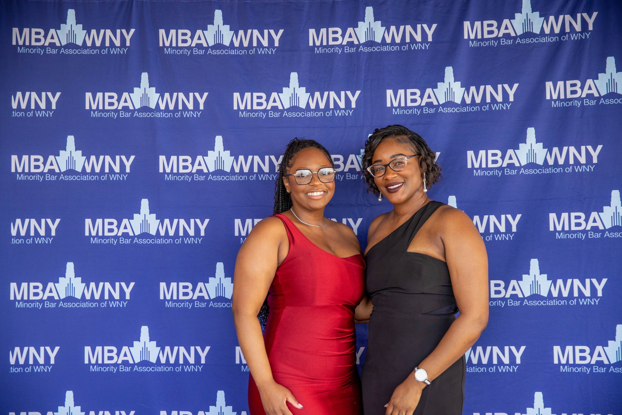 Two women in elegant dresses posing in front of a blue backdrop featuring the logos of the Minority Bar Association of WNY.