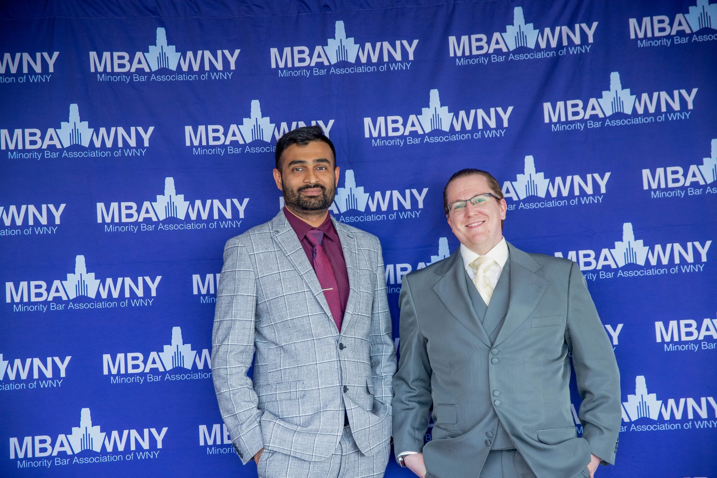 Two men dressed in suits stand in front of a blue backdrop with white logos that read "MBA WNY Minority Bar Association of WNY."
