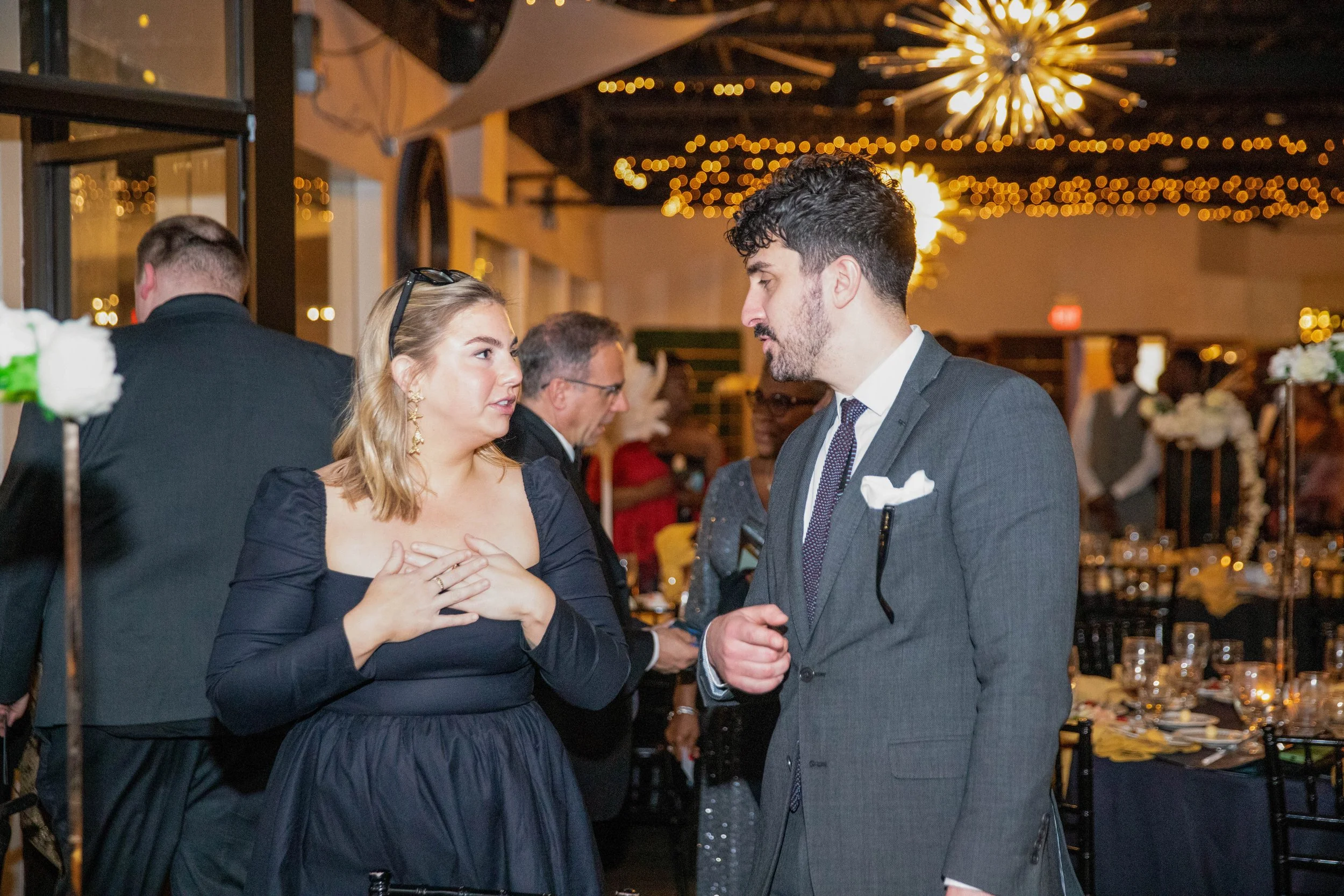 A man and woman are talking at an event, with string lights and festive decorations in the background. The woman has blonde hair, glasses on her head, and is wearing a black dress. The man has dark hair, a beard, and is wearing a gray suit with a whi