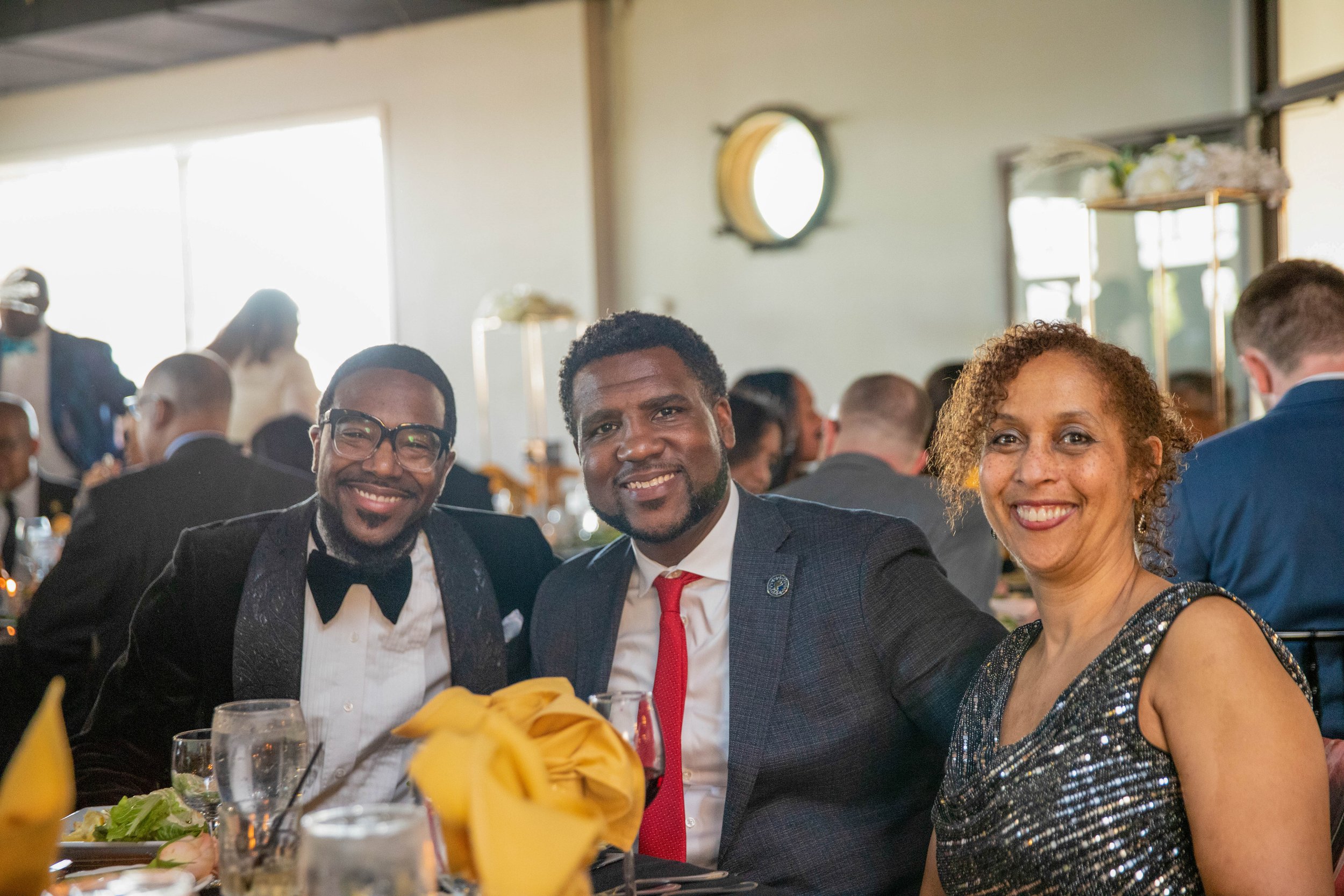Three people smiling at a formal event, seated at a table with plates, glasses, and yellow napkins, with a crowd in the background.