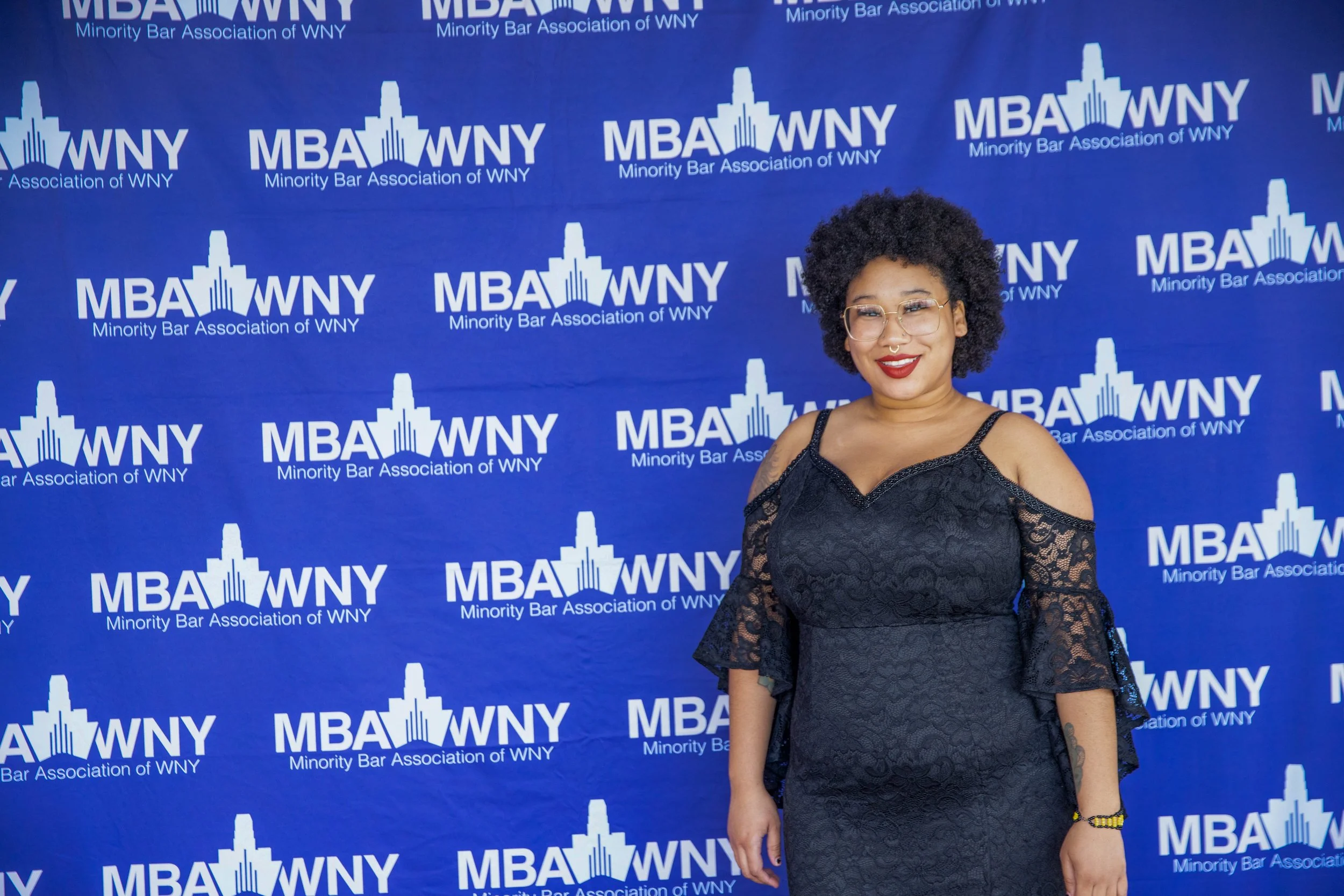 A woman standing in front of a blue backdrop with the logo and text 'MBA WNY Minority Bar Association of WNY'. She is wearing a black lace dress and glasses, smiling at the camera.