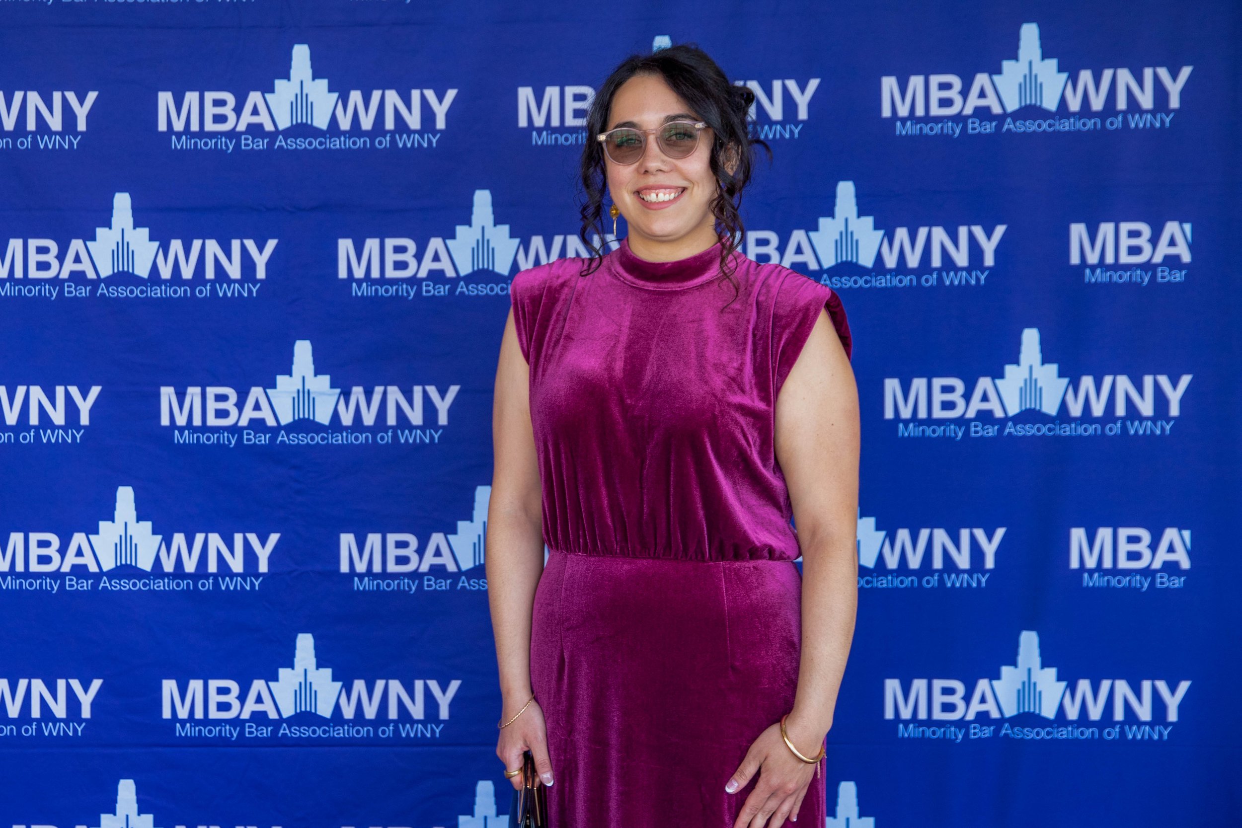 A smiling woman in a purple velvet dress standing in front of a blue backdrop with the logo and name of the Minority Bar Association of WNY.