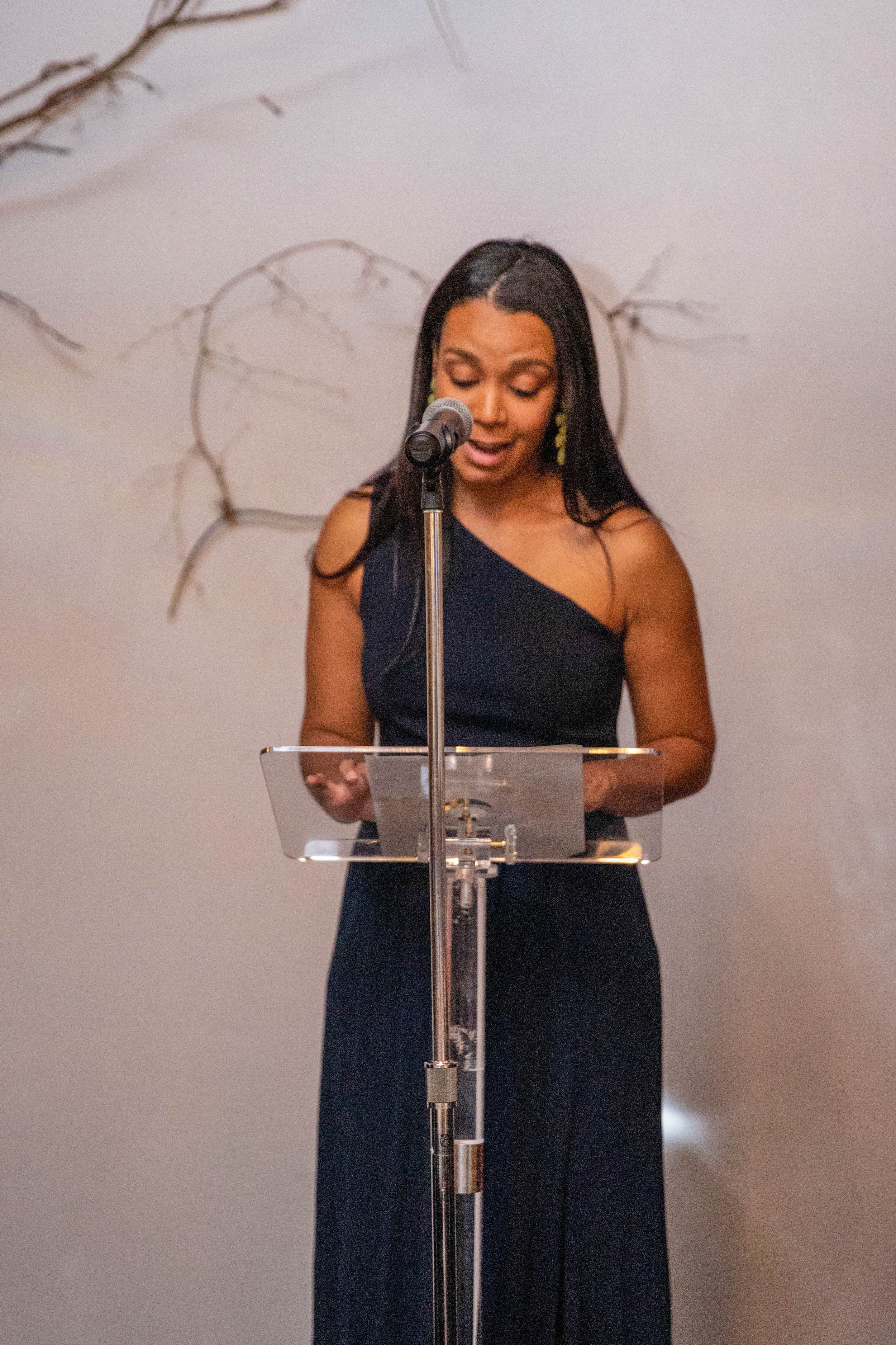 A woman in a black dress stands at a clear lectern, reading or speaking into a microphone, with a minimalist white wall behind her decorated with a sparse, artistic branch design.