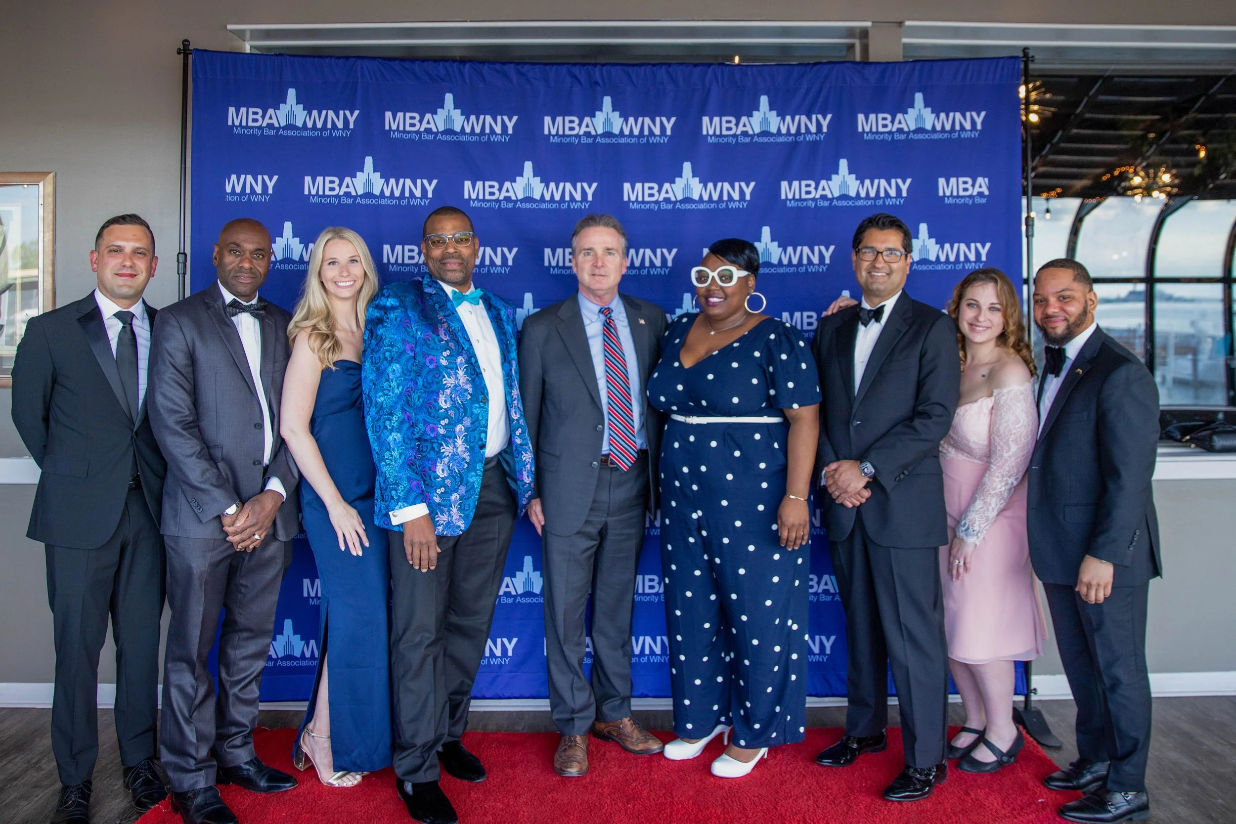 A diverse group of nine people dressed in formal attire posing for a photo on a red carpet in front of a blue backdrop with the logo and text of the Minority Bar Association of WNY.
