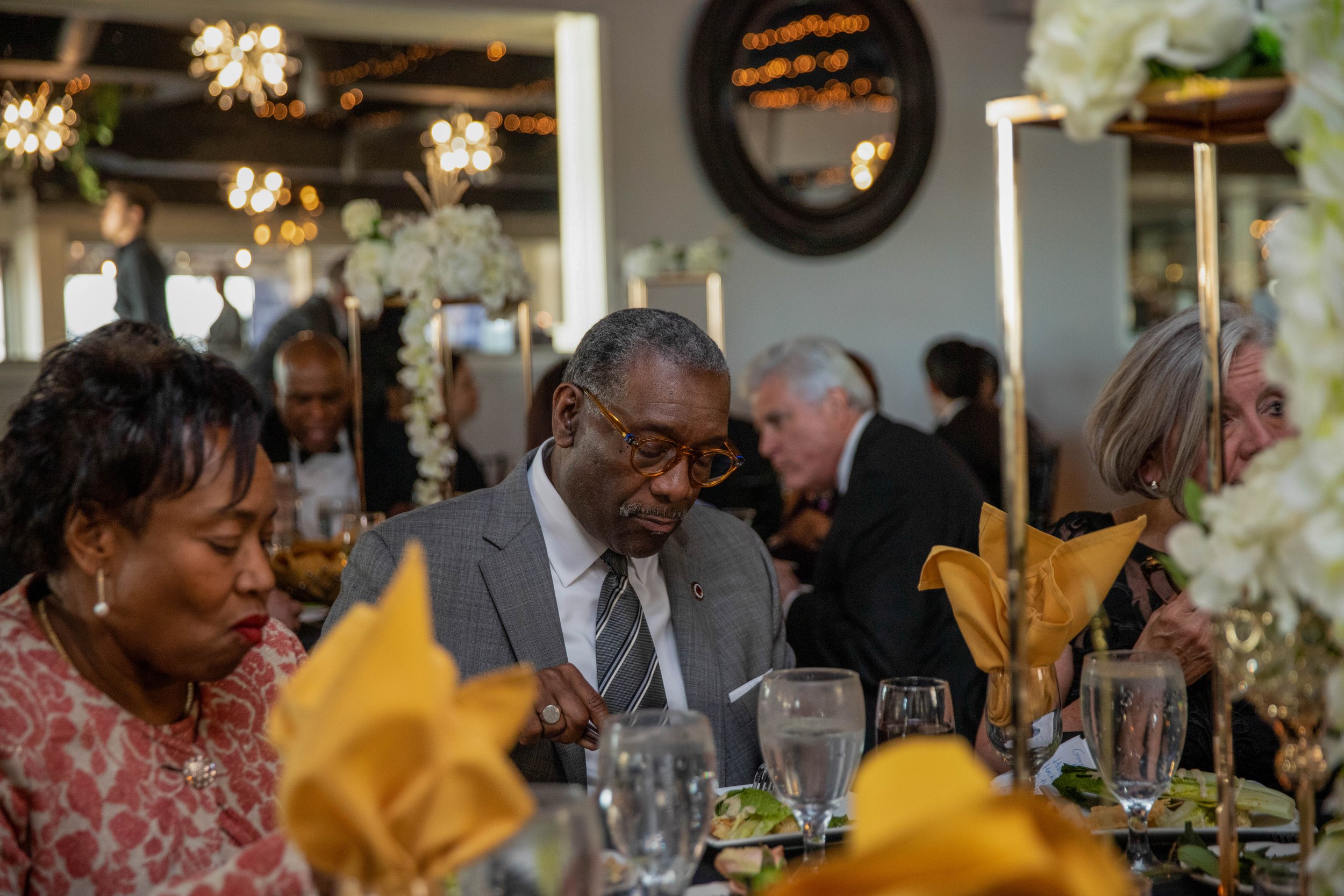 People seated at a formal event decorated with floral arrangements and candlelit chandeliers.