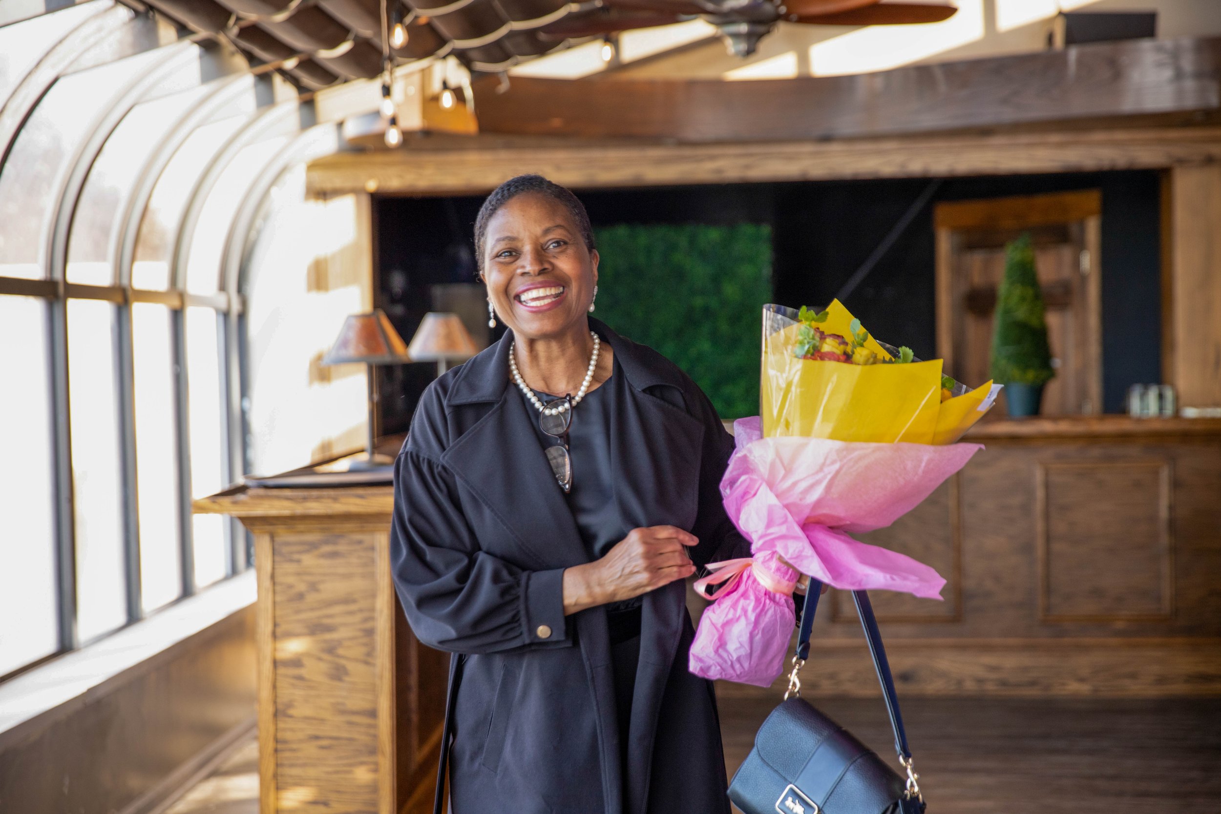 A smiling woman holding a bouquet of flowers in a pink wrapping paper, wearing a black jacket and pearl jewelry inside a room with wooden decor and large windows.
