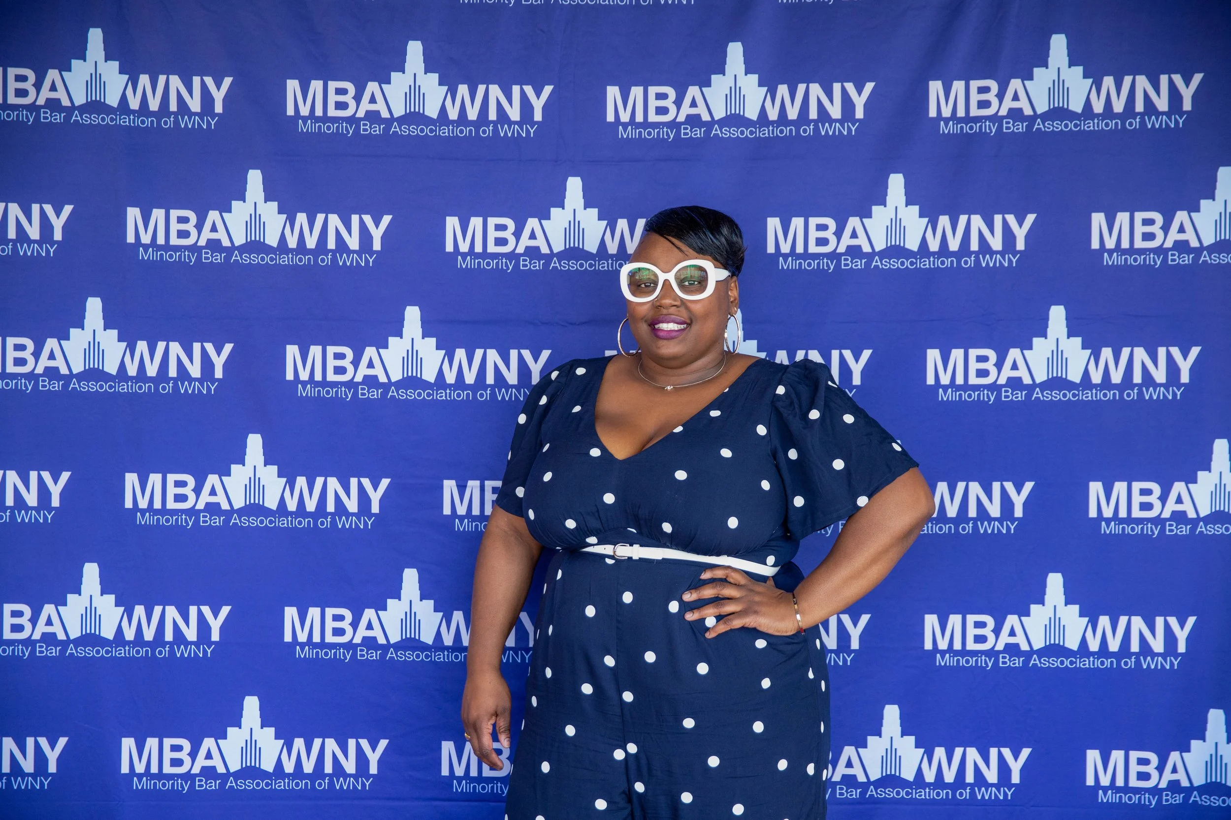 A woman wearing sunglasses and hoop earrings, dressed in a navy blue dress with white polka dots, standing in front of a backdrop with the logo and text of the Minority Bar Association of WNY and MBA WNY.