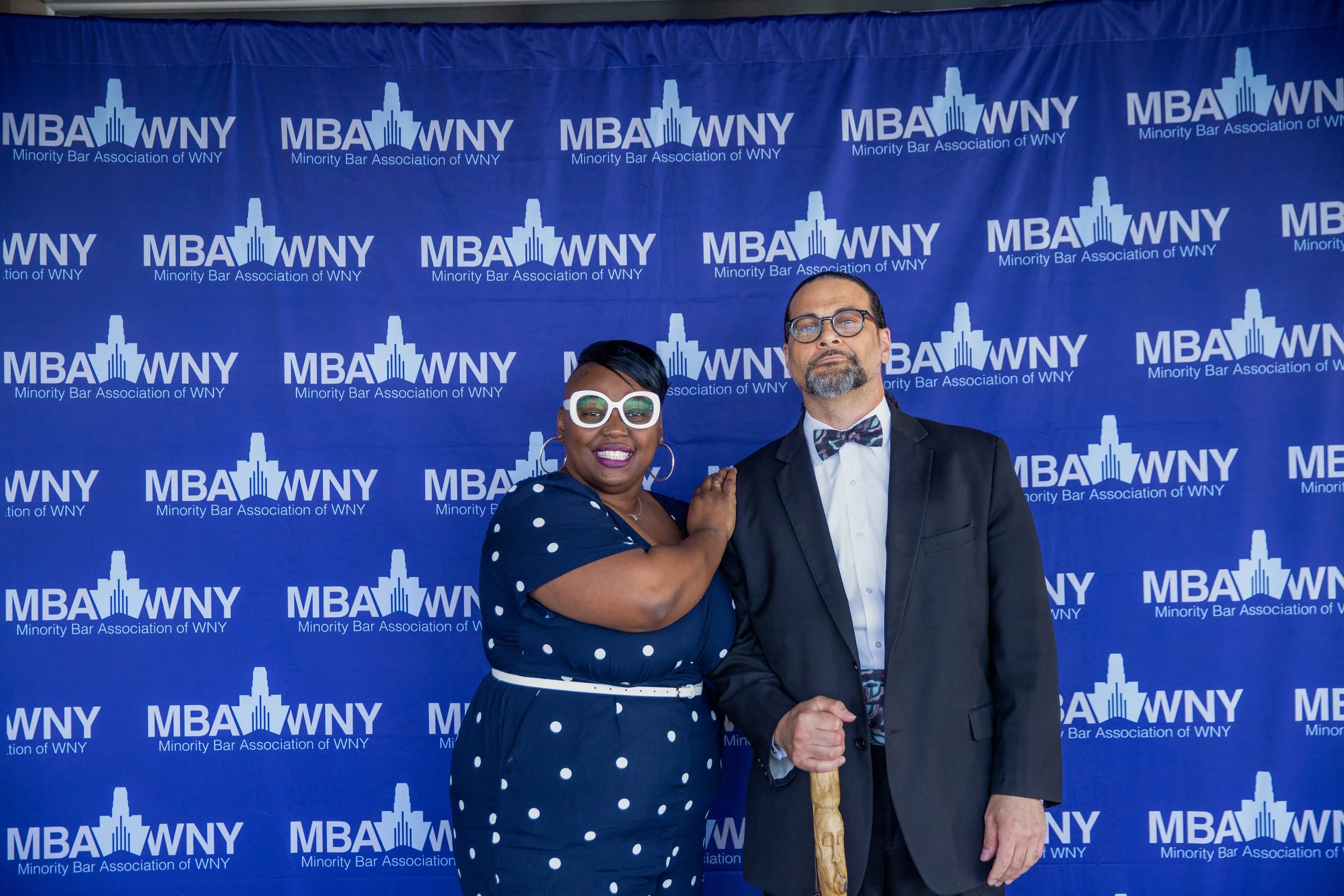 Two individuals standing in front of a blue backdrop that displays the logo of the Minority Bar Association of WNY. The woman on the left is wearing a navy blue polka dot dress with glasses and large earrings, and is smiling while holding the man's a