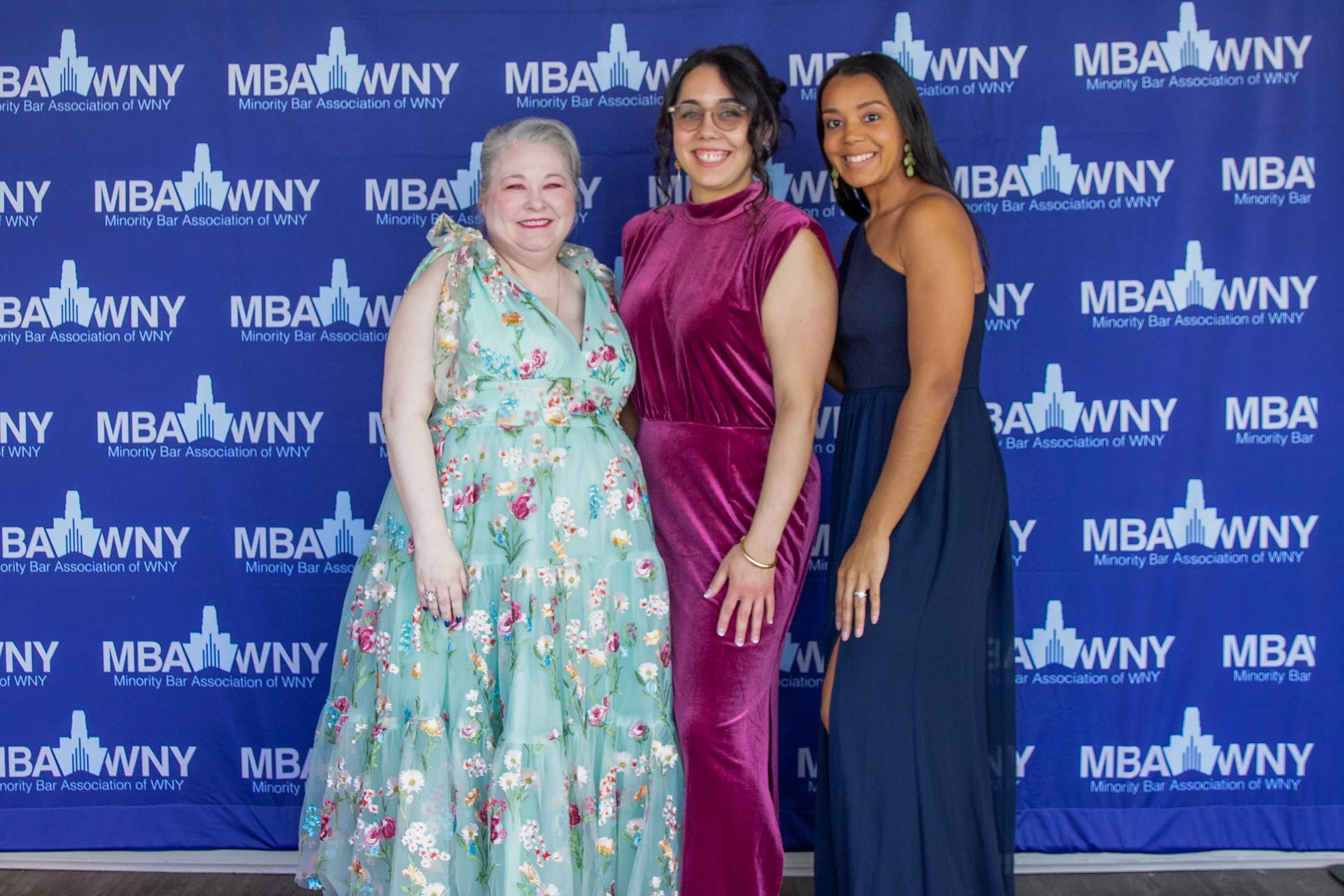 Three women standing in front of a blue backdrop with 'MBA WNY Minority Bar Association of WNY' logo, smiling and dressed in colorful formal attire.