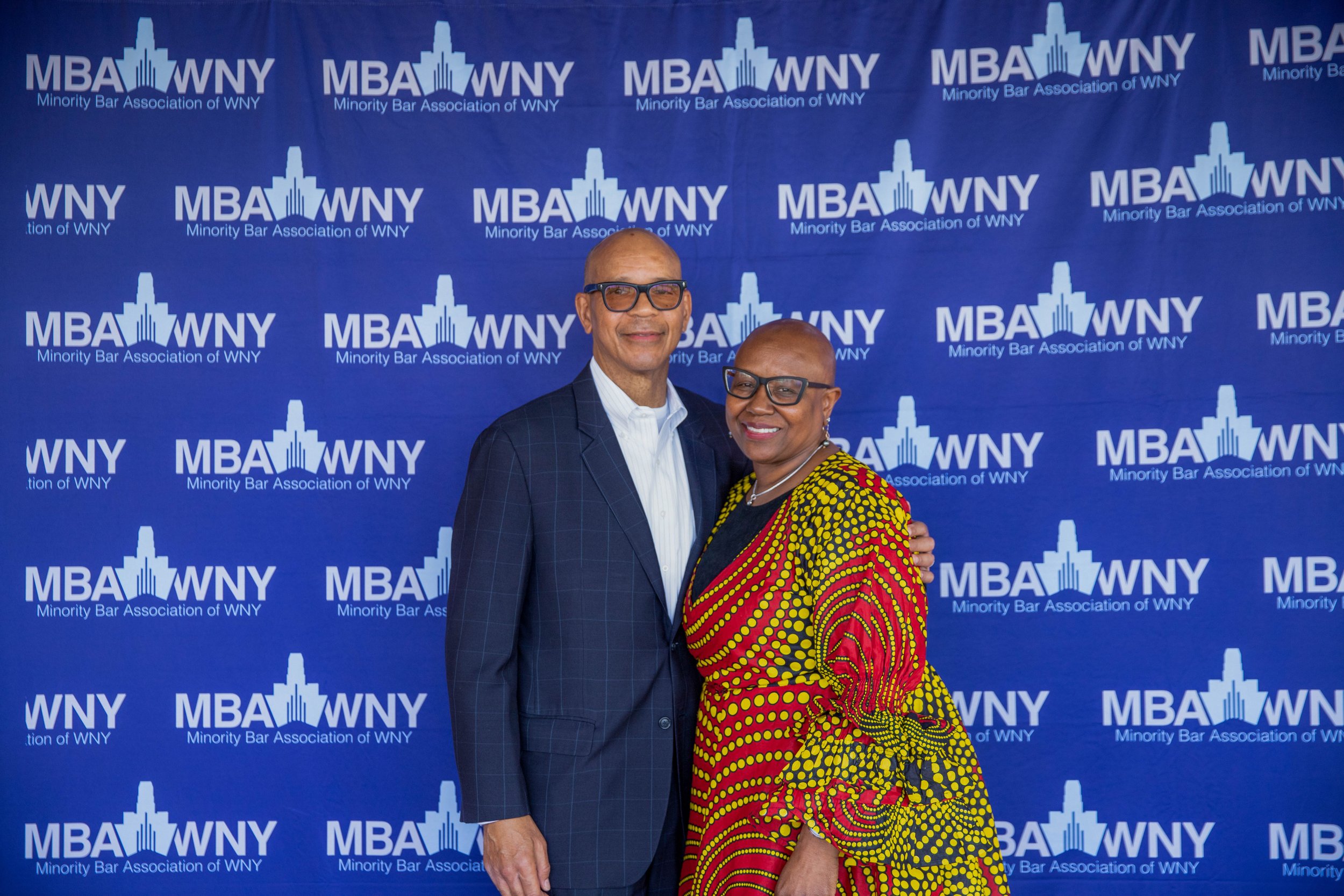 Two individuals, a man and a woman, standing together at an event with a backdrop displaying the logo and name of the Minority Bar Association of WNY. The man is dressed in a checkered blazer and glasses, and the woman is wearing glasses and a colorf